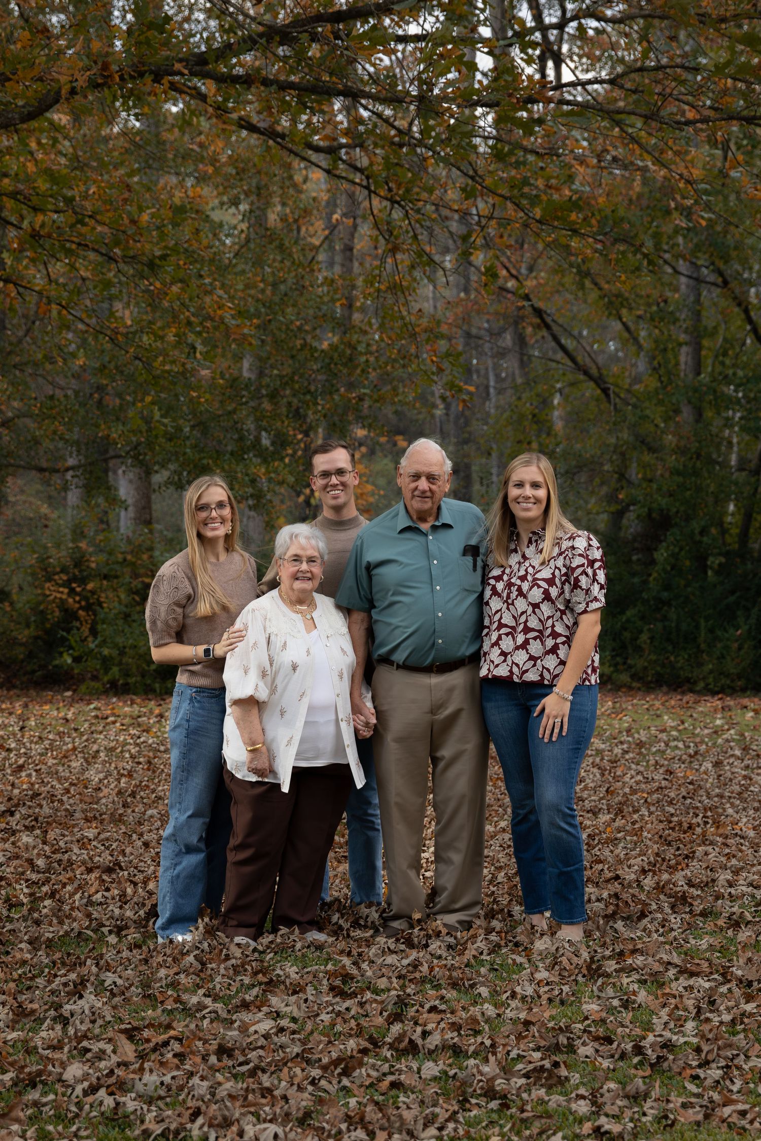 Family of five posing outdoors among autumn leaves with colorful fall foliage trees in background.