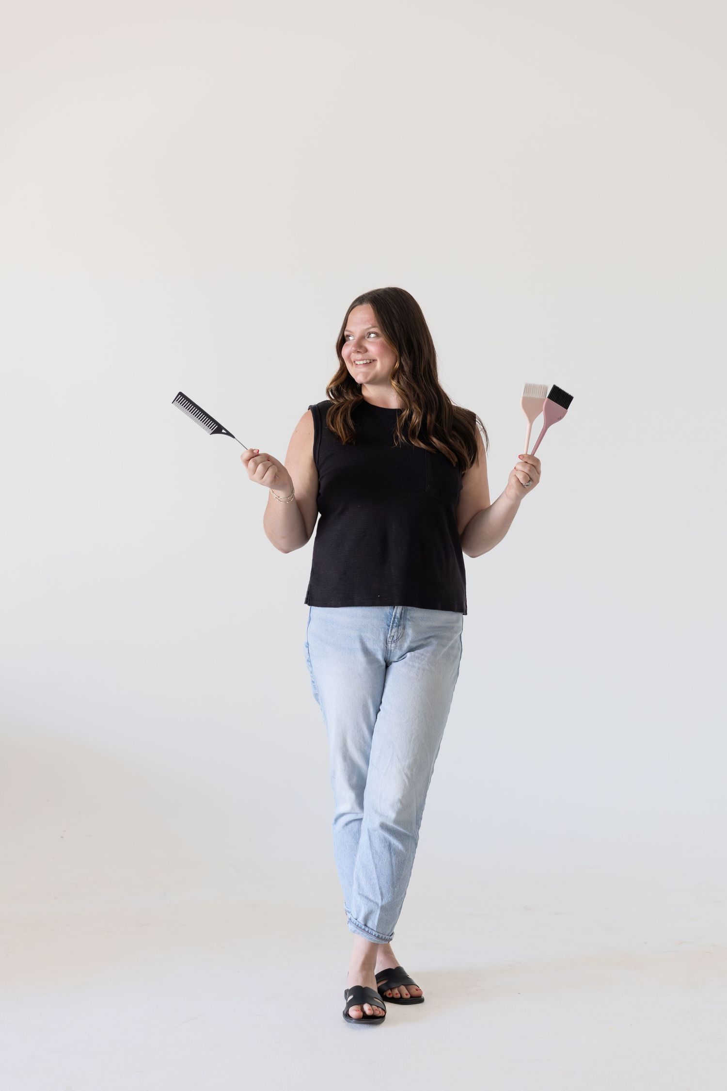Happy woman in black sleeveless top and jeans posing with kitchen utensils on white background.
