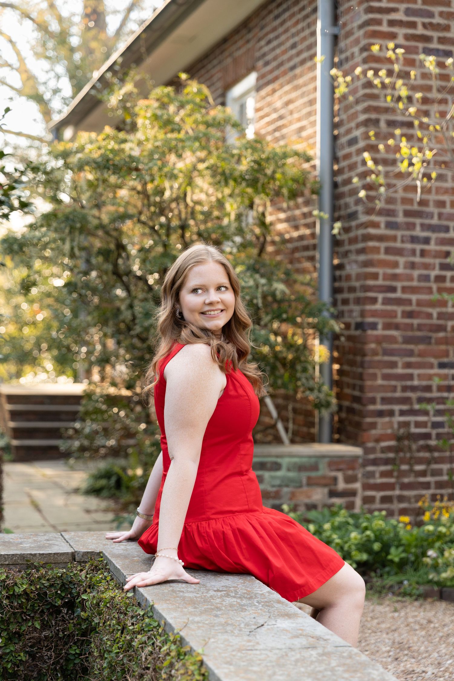 Smiling young woman in red dress sitting on stone wall outside brick building.