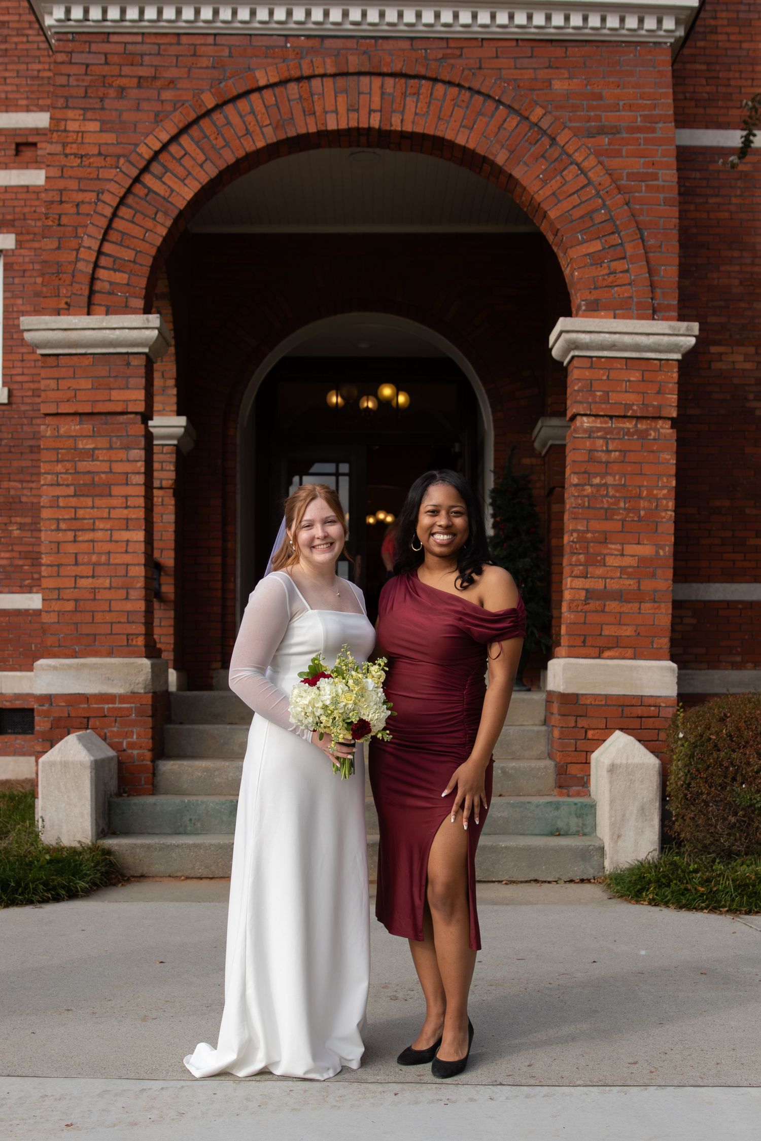 Bride holding bouquet in white gown posing with bridesmaid in burgundy dress outside red brick venue.