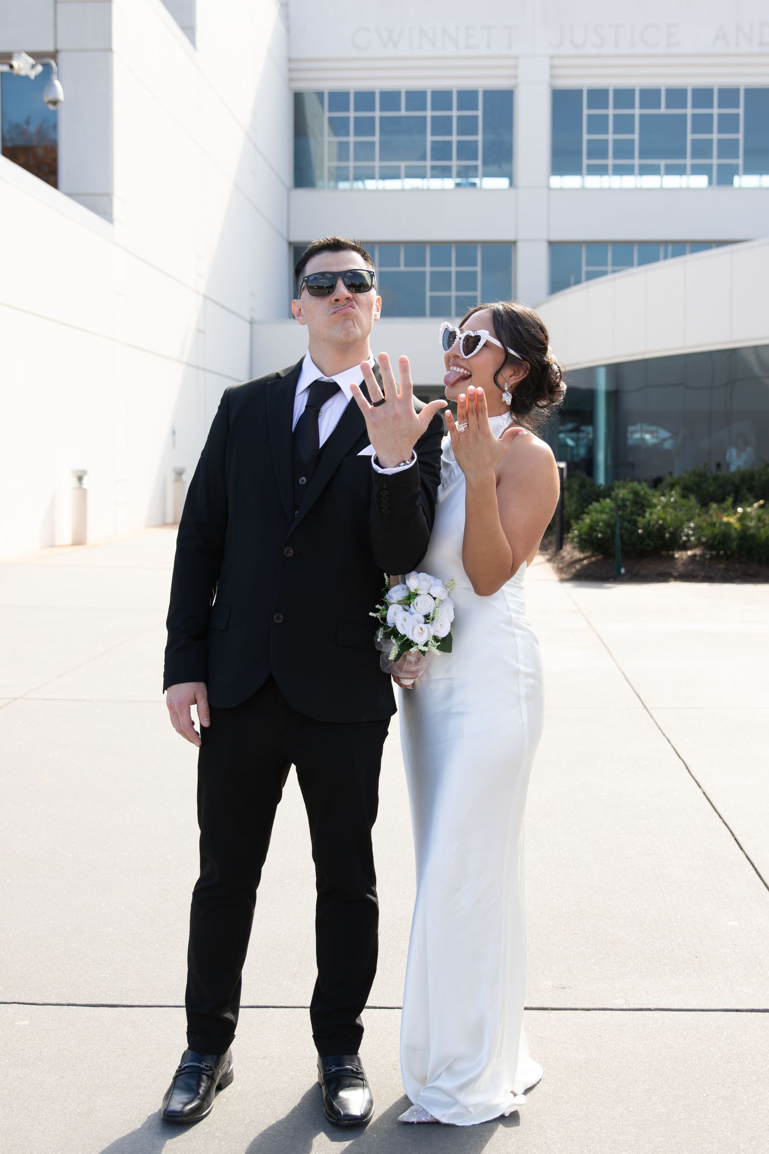 Newlywed couple in sunglasses showing wedding rings outside, groom in black suit, bride in white gown holding bouquet.
