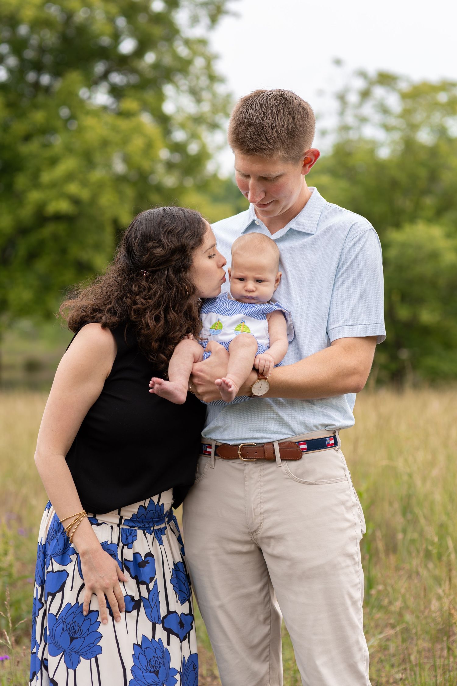 Mother kissing newborn baby held by father during outdoor family photo session in green field.