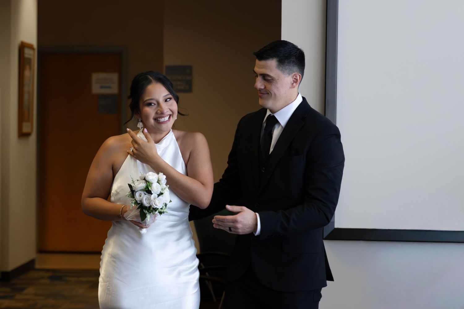 Bride in white halter gown holding bouquet laughs beside groom in black suit during wedding ceremony.