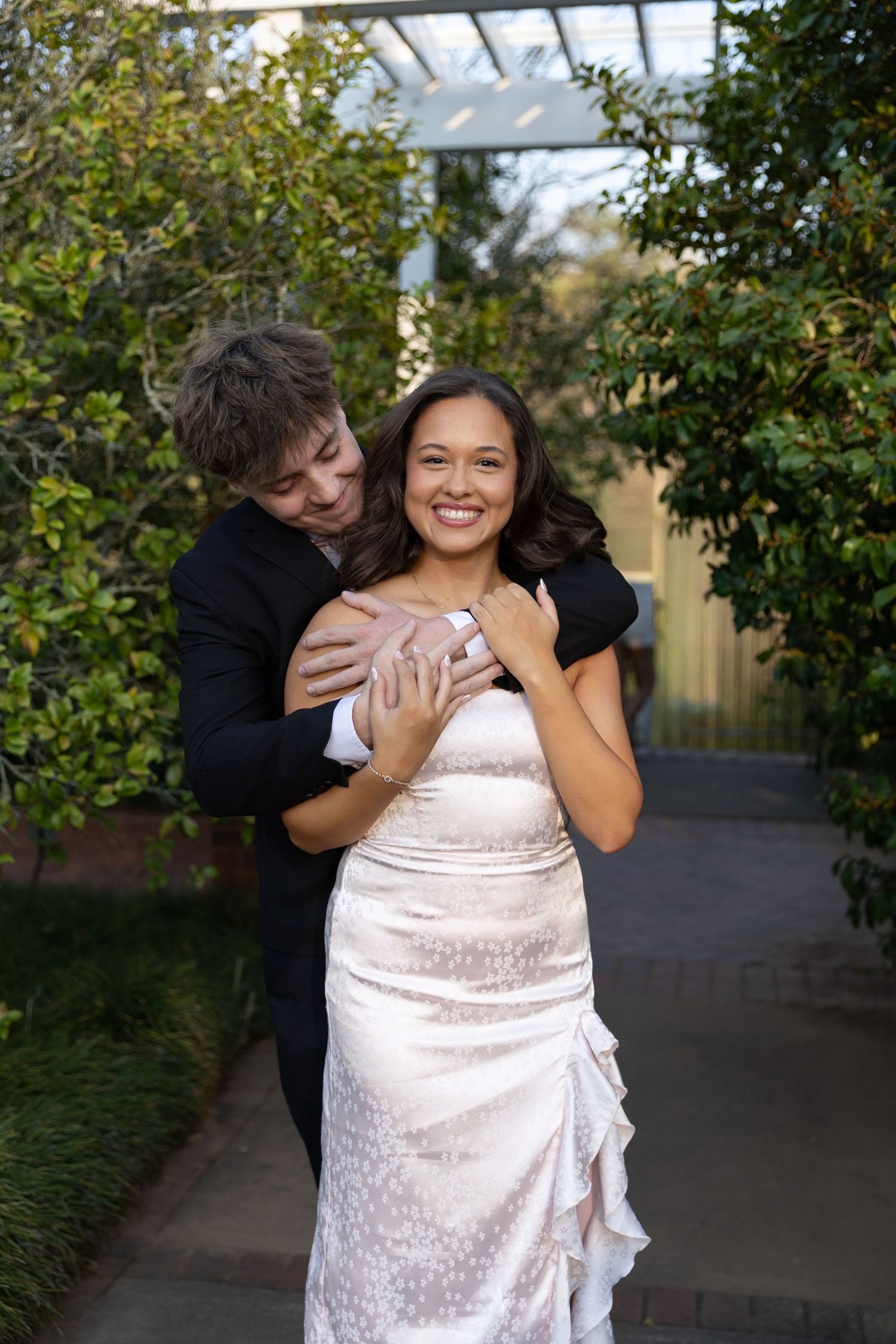 Couple in formal wear embracing outdoors, bride in white gown smiling, groom in black suit, garden background.