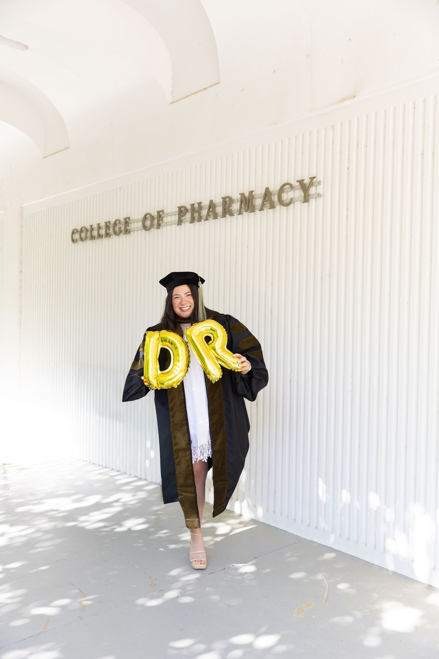 Pharmacy graduate in cap and gown holding gold DR balloons at College of Pharmacy.