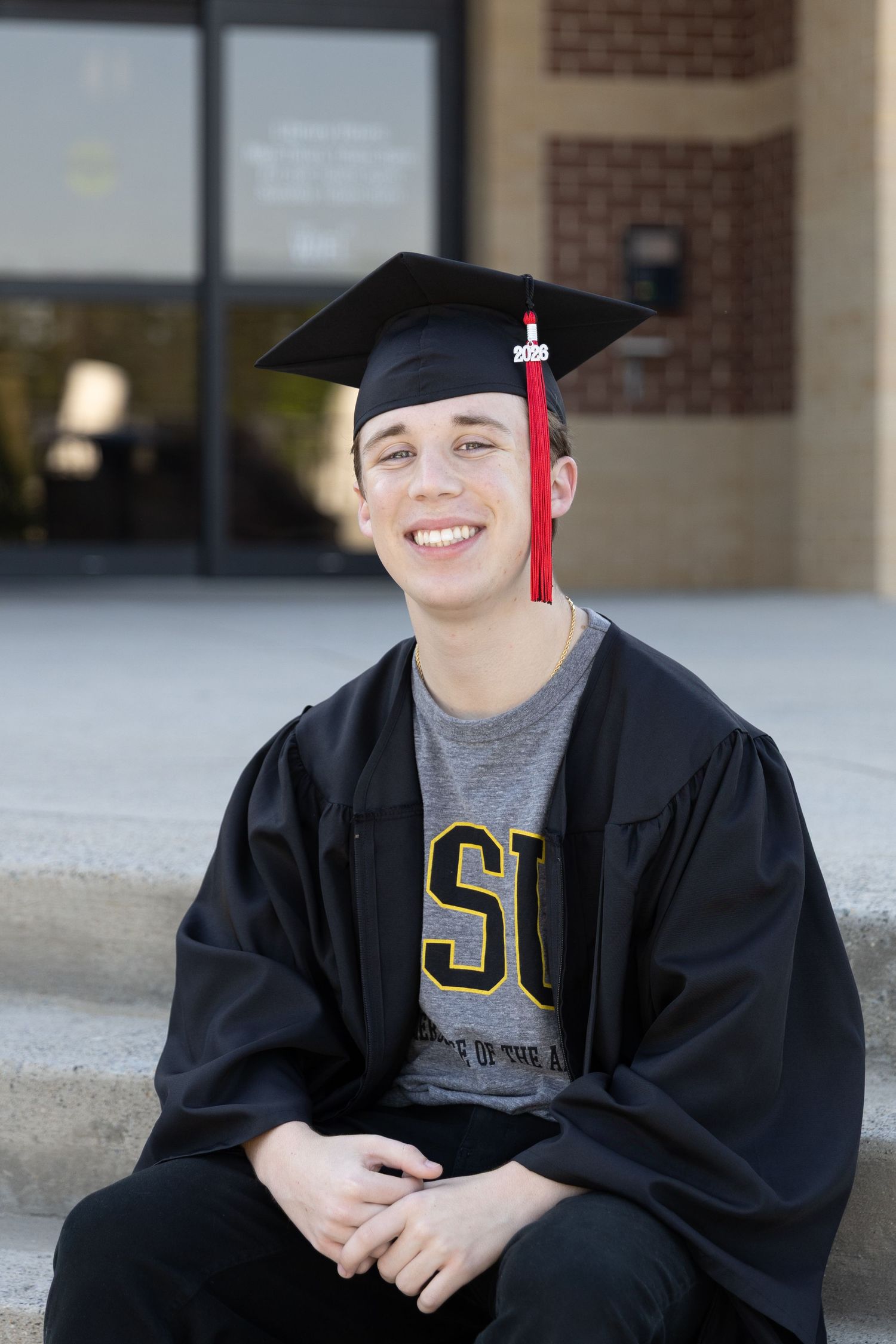 Smiling young man in black graduation cap and gown with red tassel sits on steps wearing an JSU college shirt.