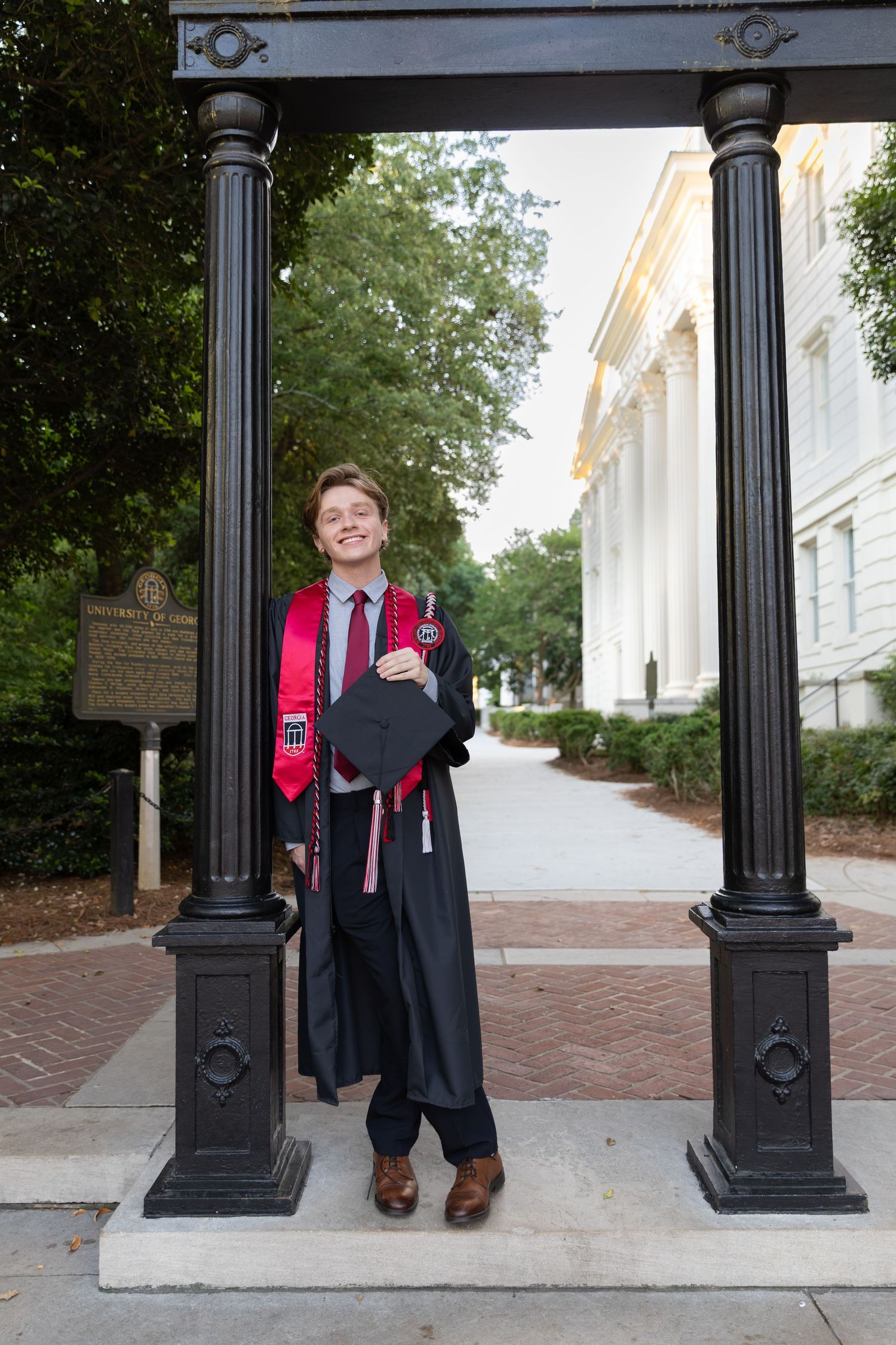 A smiling graduate in black gown and red stole holds cap between two columns on a brick university campus pathway.