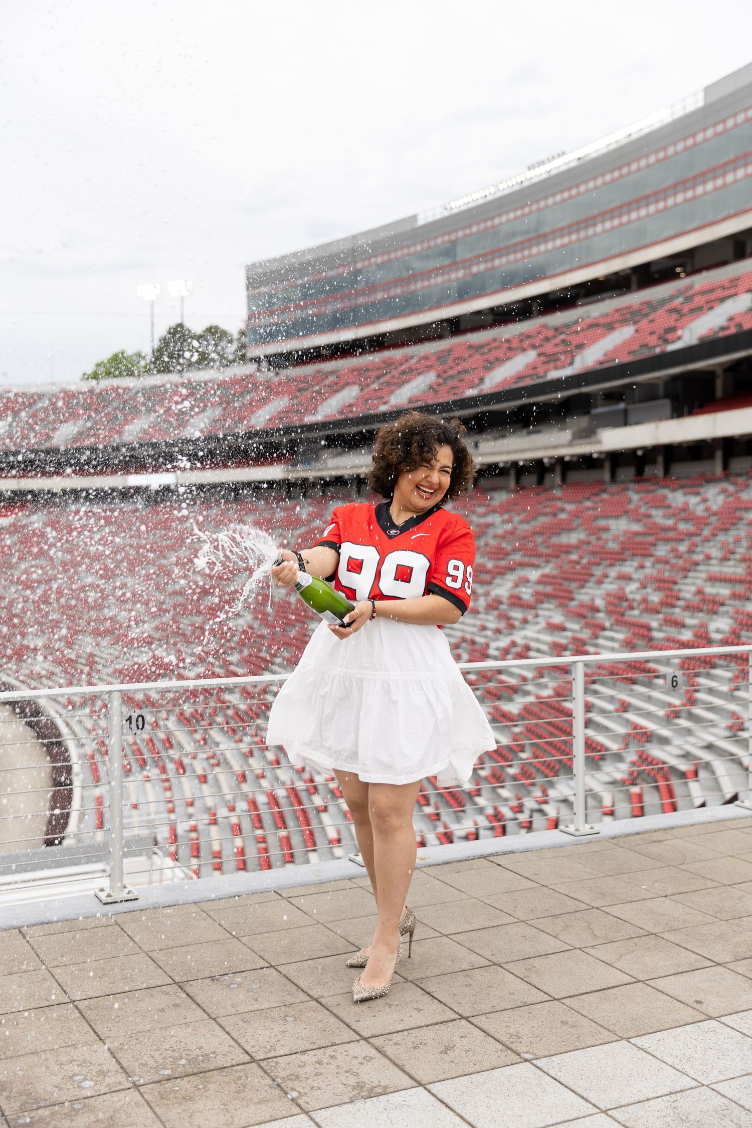 Woman in red football jersey and white skirt popping champagne bottle at a large stadium.