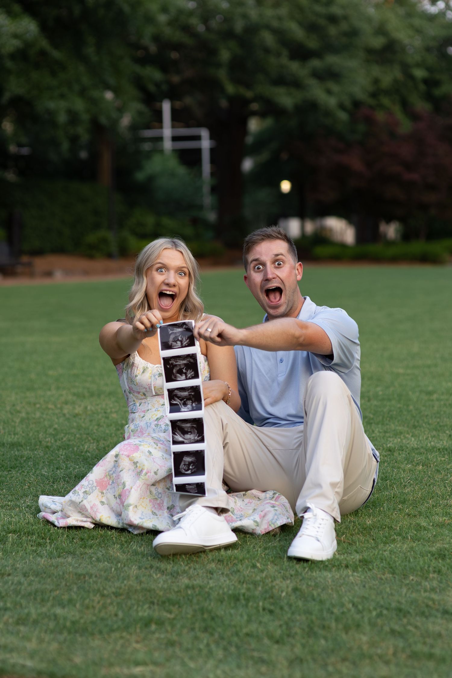 Excited couple sits on grass with open mouths holding ultrasound images in surprise.