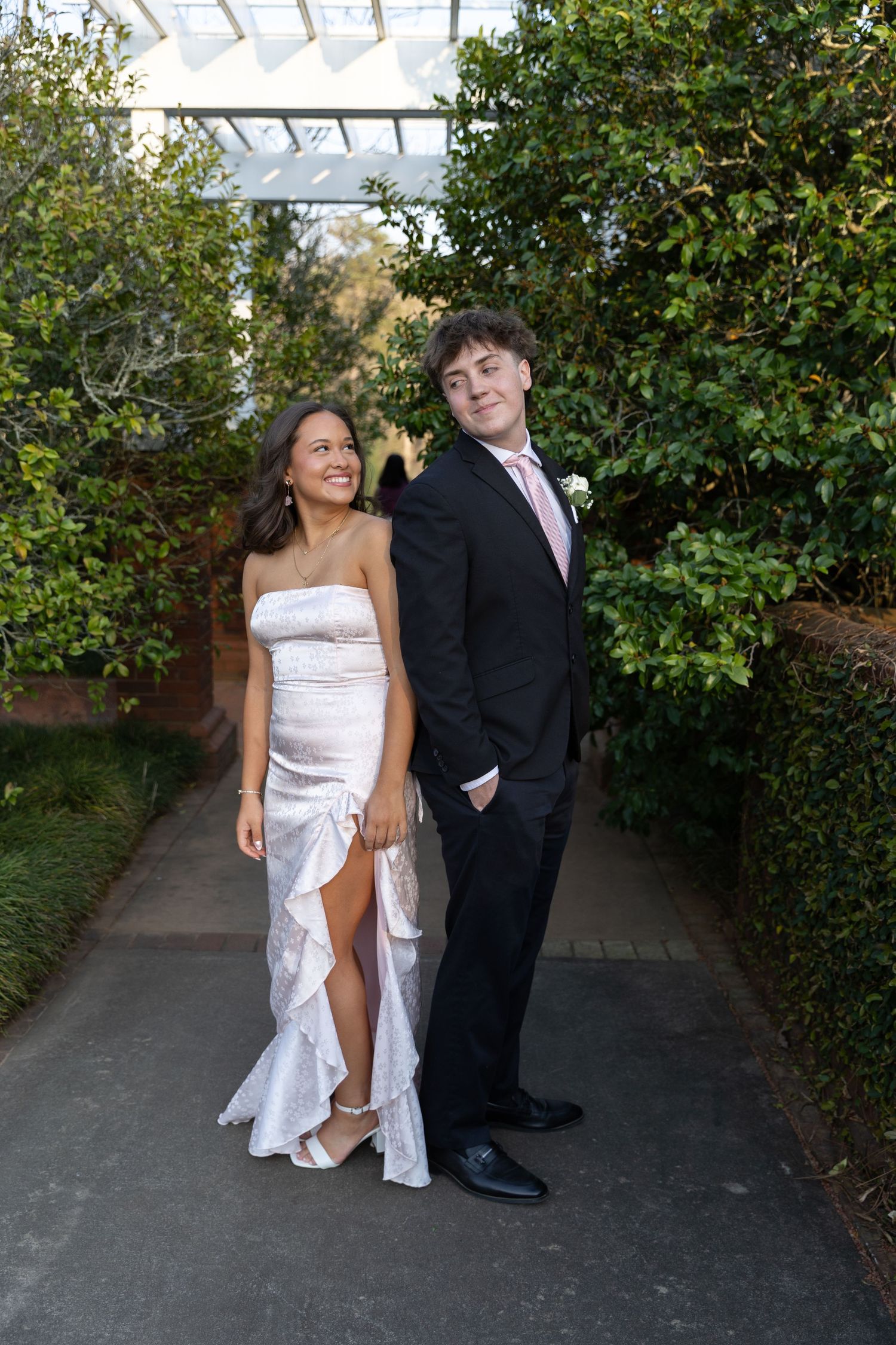 A young couple dressed in formal prom attire pose together in a garden pathway, she in white dress, he in black suit.
