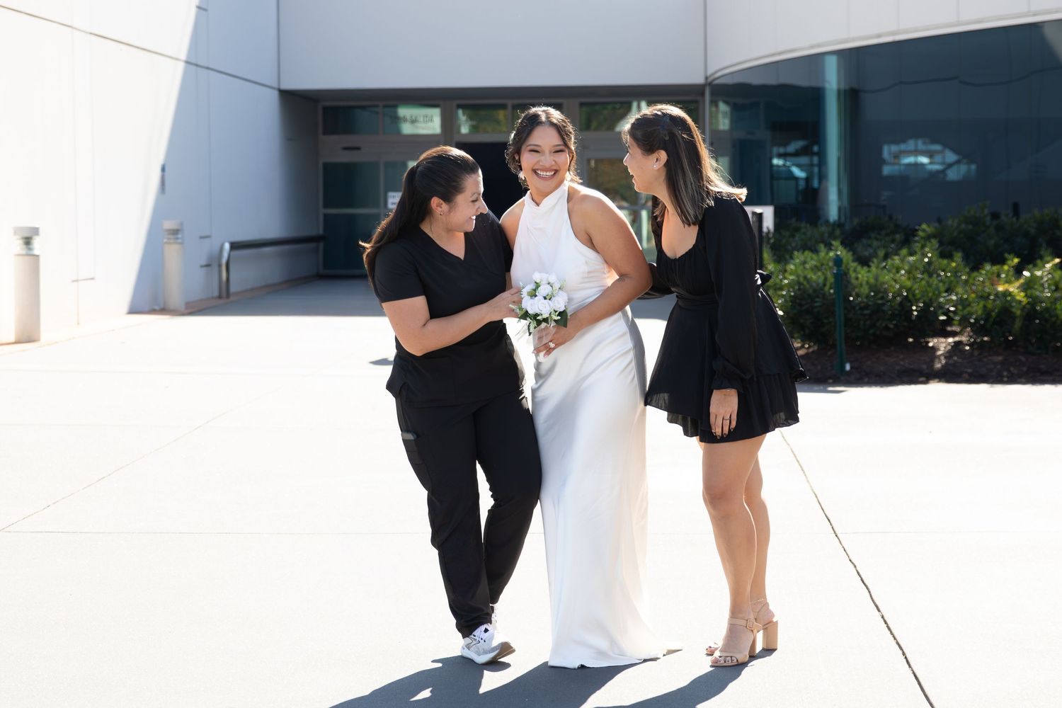 Bride in white gown holding bouquet flanked by two women in black outfits outdoors on sunny day.