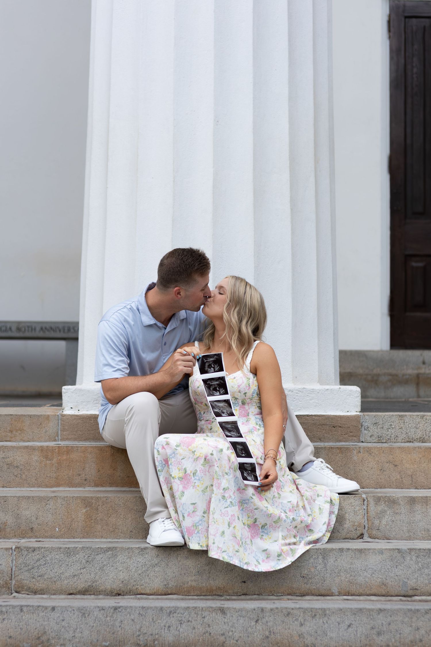 Couple kissing on stone steps holding ultrasound photos in a floral dress pregnancy announcement.