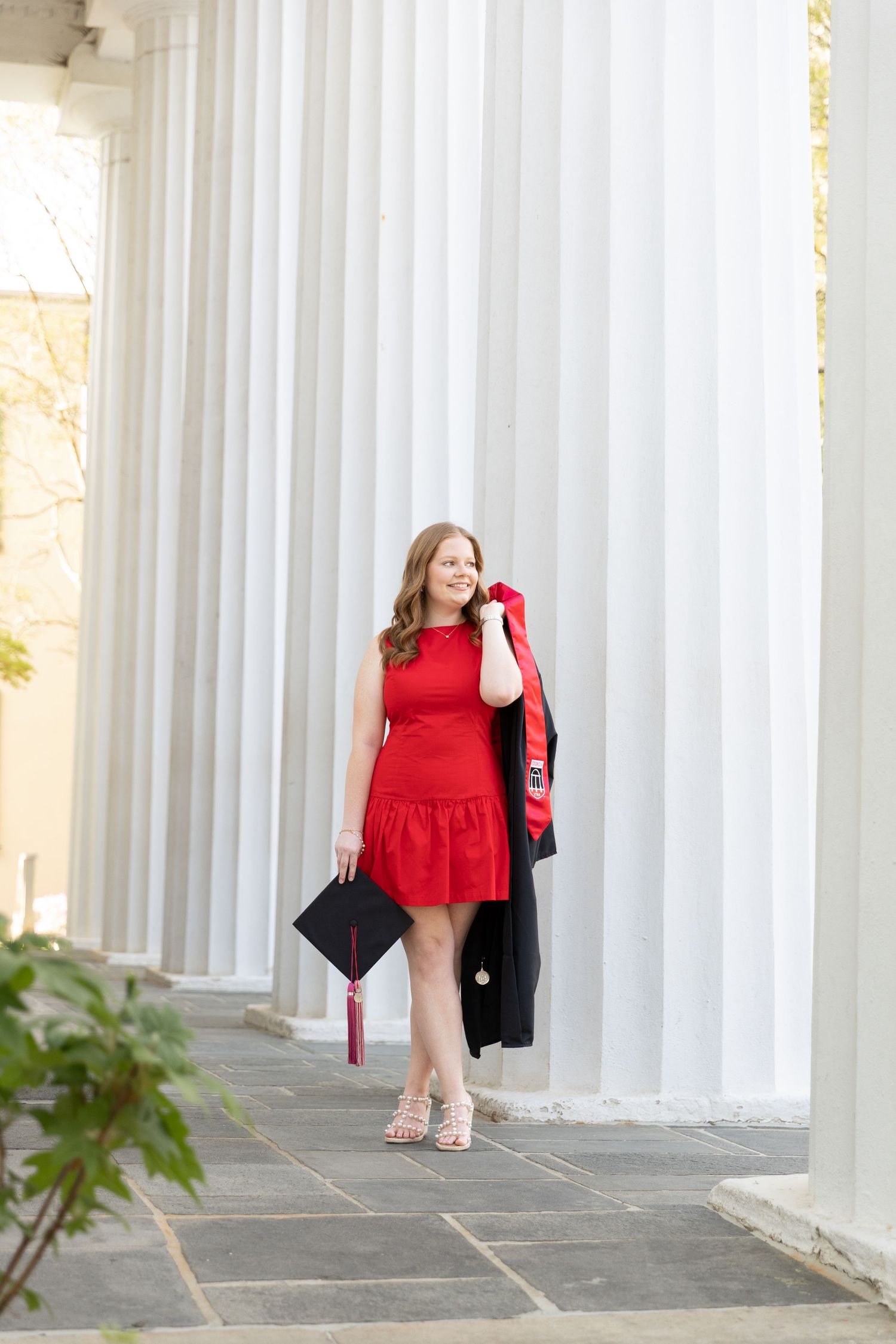 Young woman in red dress holding graduation cap and gown, posing between white columns for senior photos.