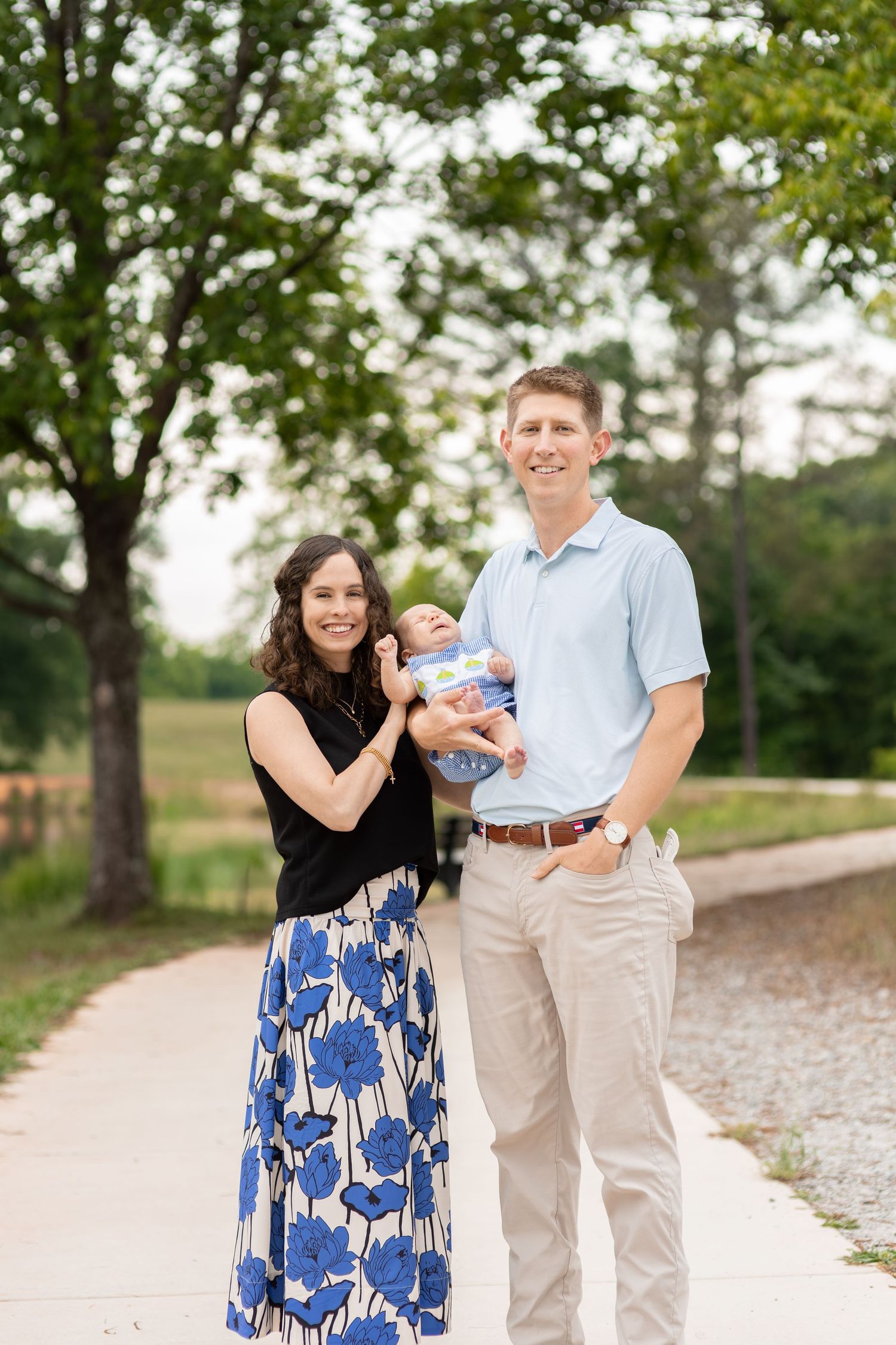 A smiling couple stands outdoors on a path holding their newborn baby, woman in floral skirt and man in light blue polo.