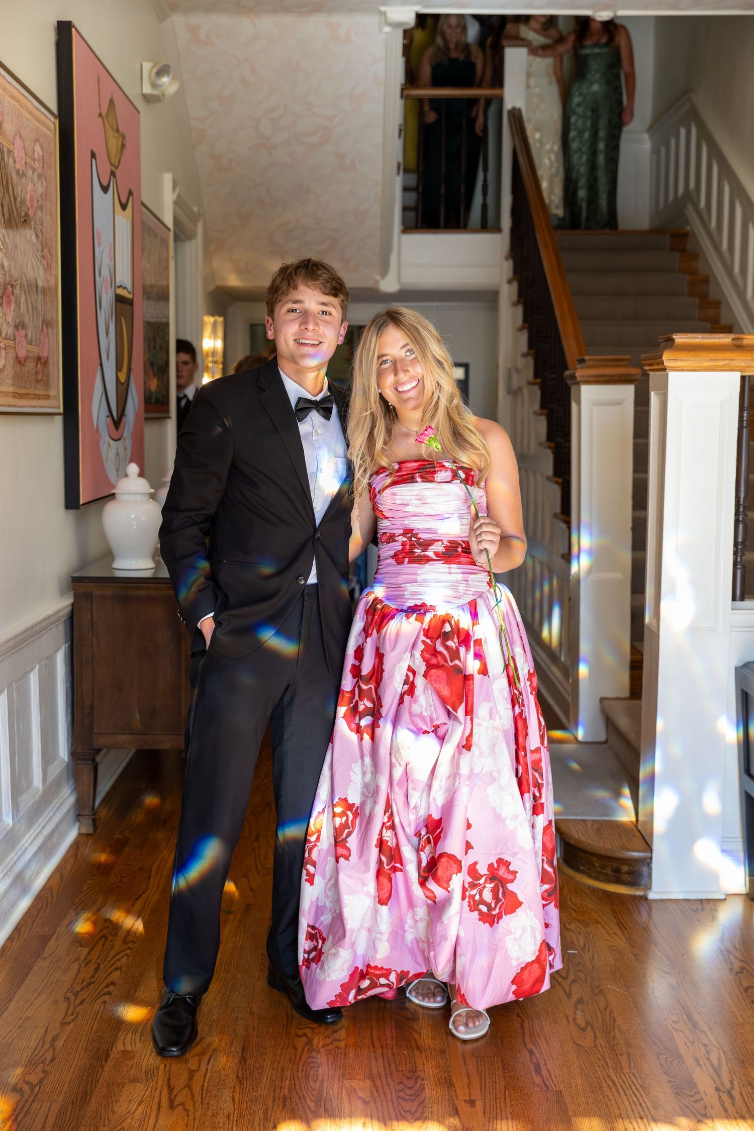Young couple in formal prom attire posing indoors, man in tuxedo and woman in floral gown.