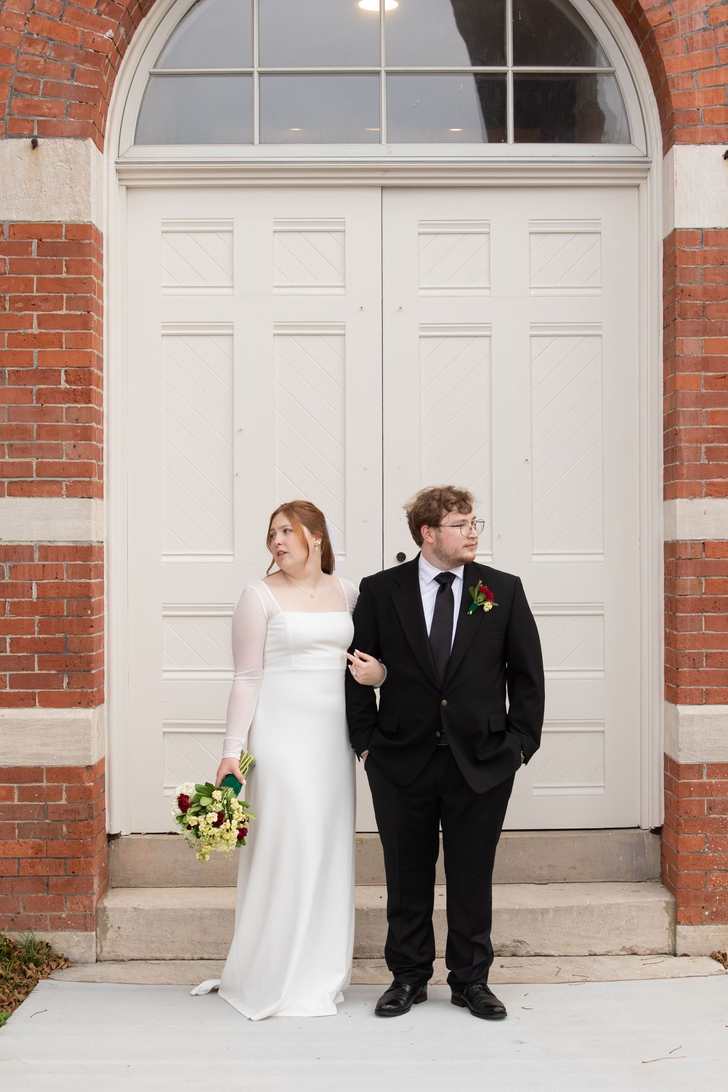 Bride and groom stand before elegant white double doors of classic red brick building on wedding day.