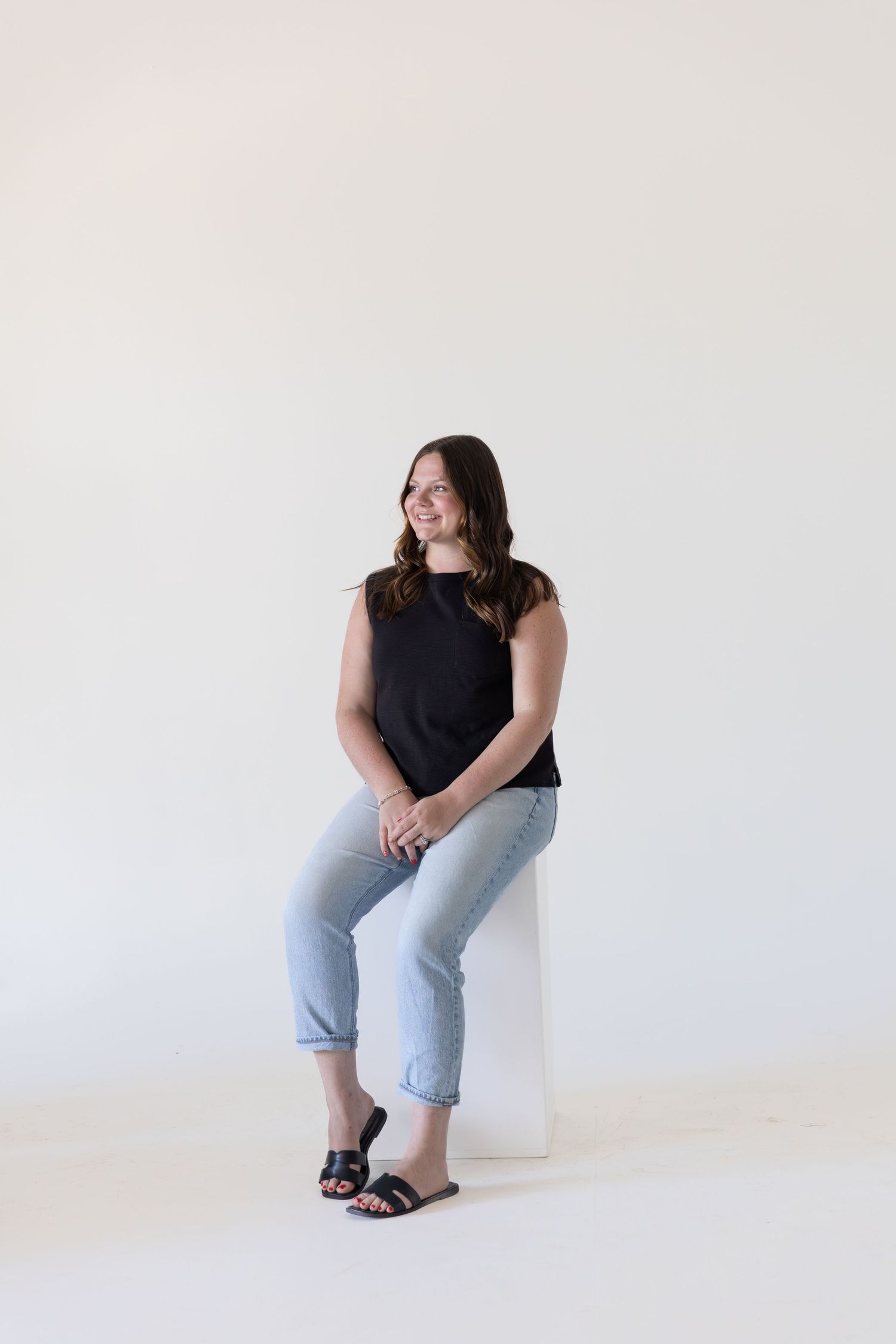Woman in black top and light jeans sitting on white block, smiling, against a clean white background.