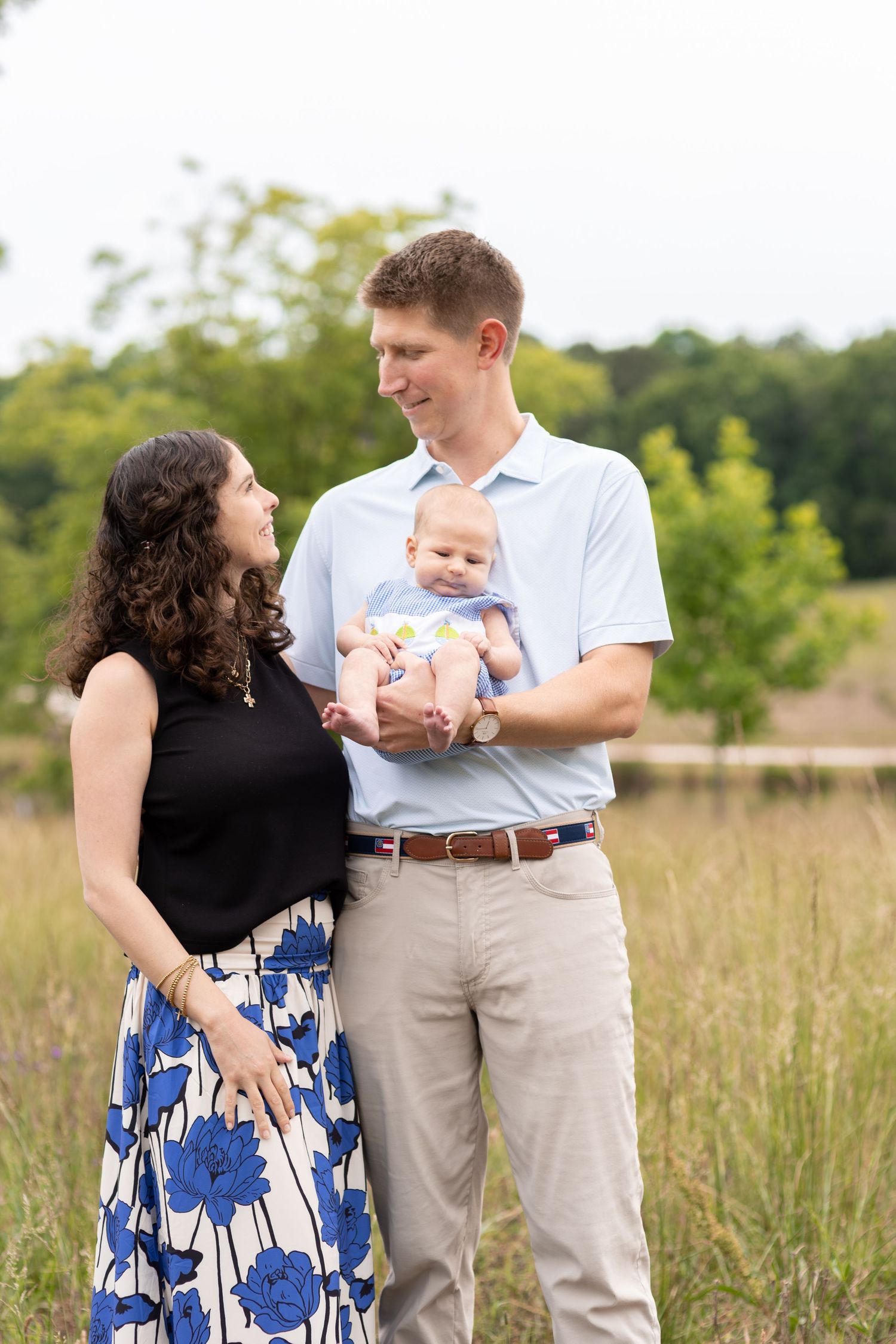 Young couple smiling at each other while holding their newborn baby in an outdoor field setting.