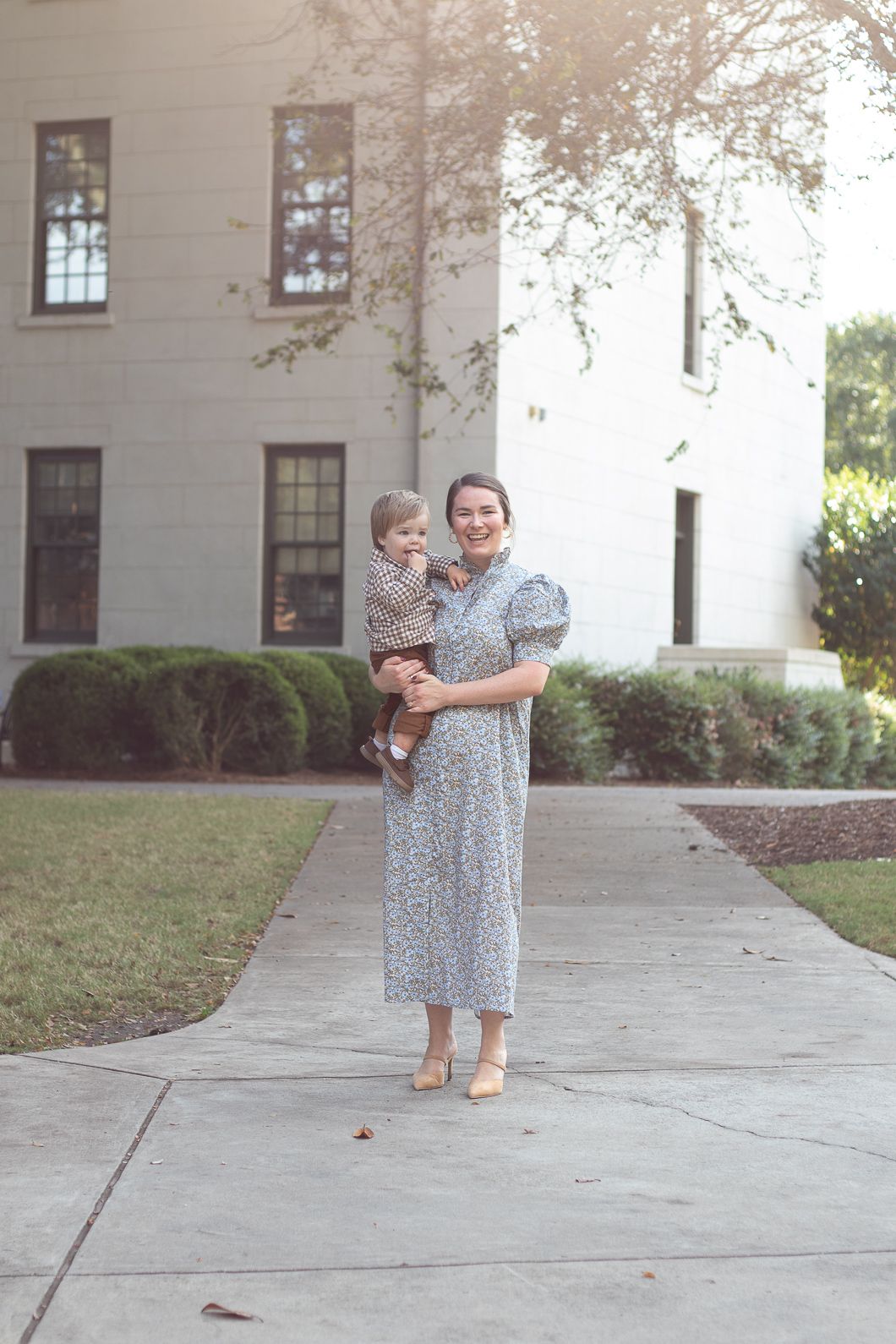 Woman in floral dress holds toddler on a sunny sidewalk outside a white building.