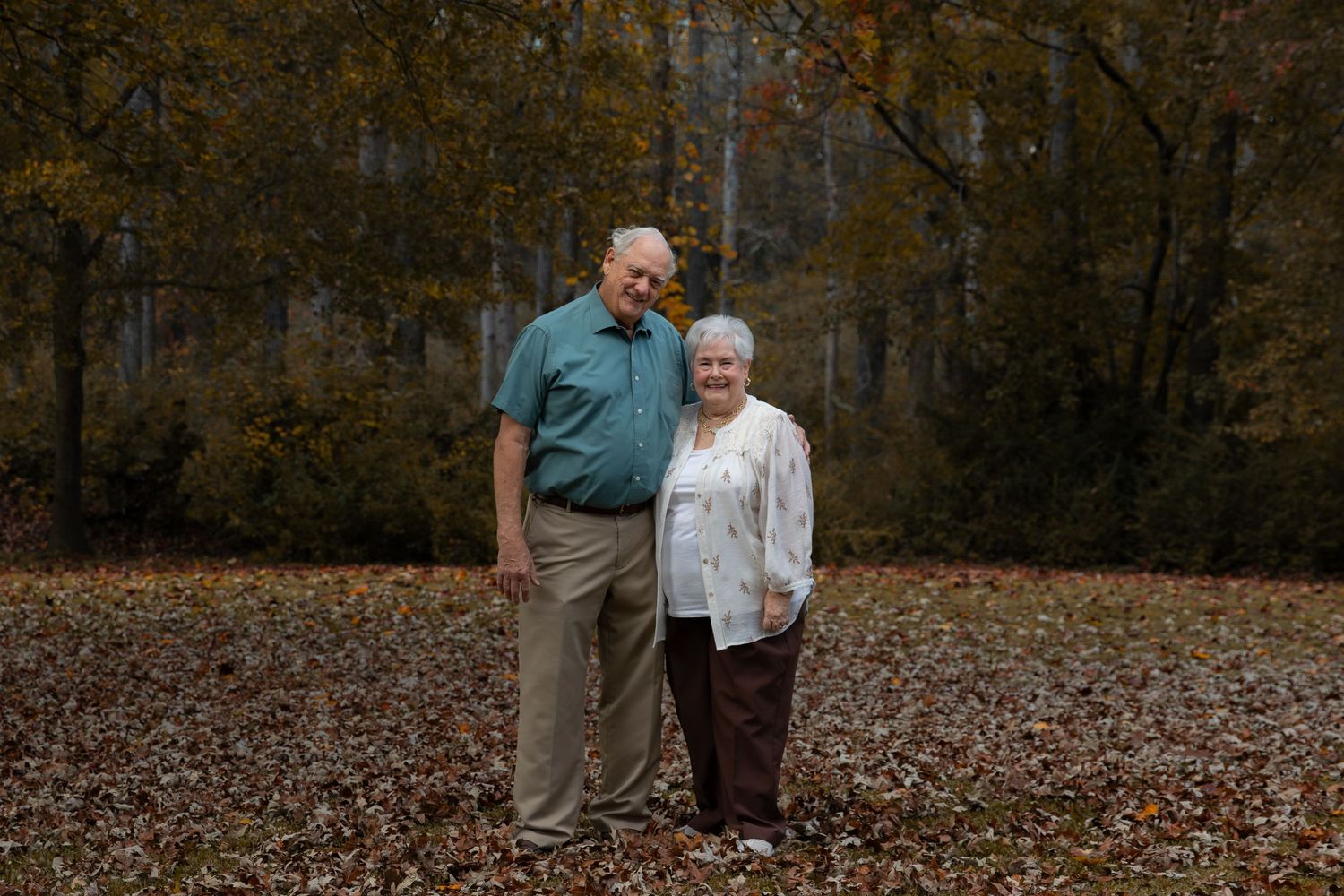 Elderly couple smiling together outdoors in autumn, surrounded by fallen leaves and colorful trees in a wooded setting.