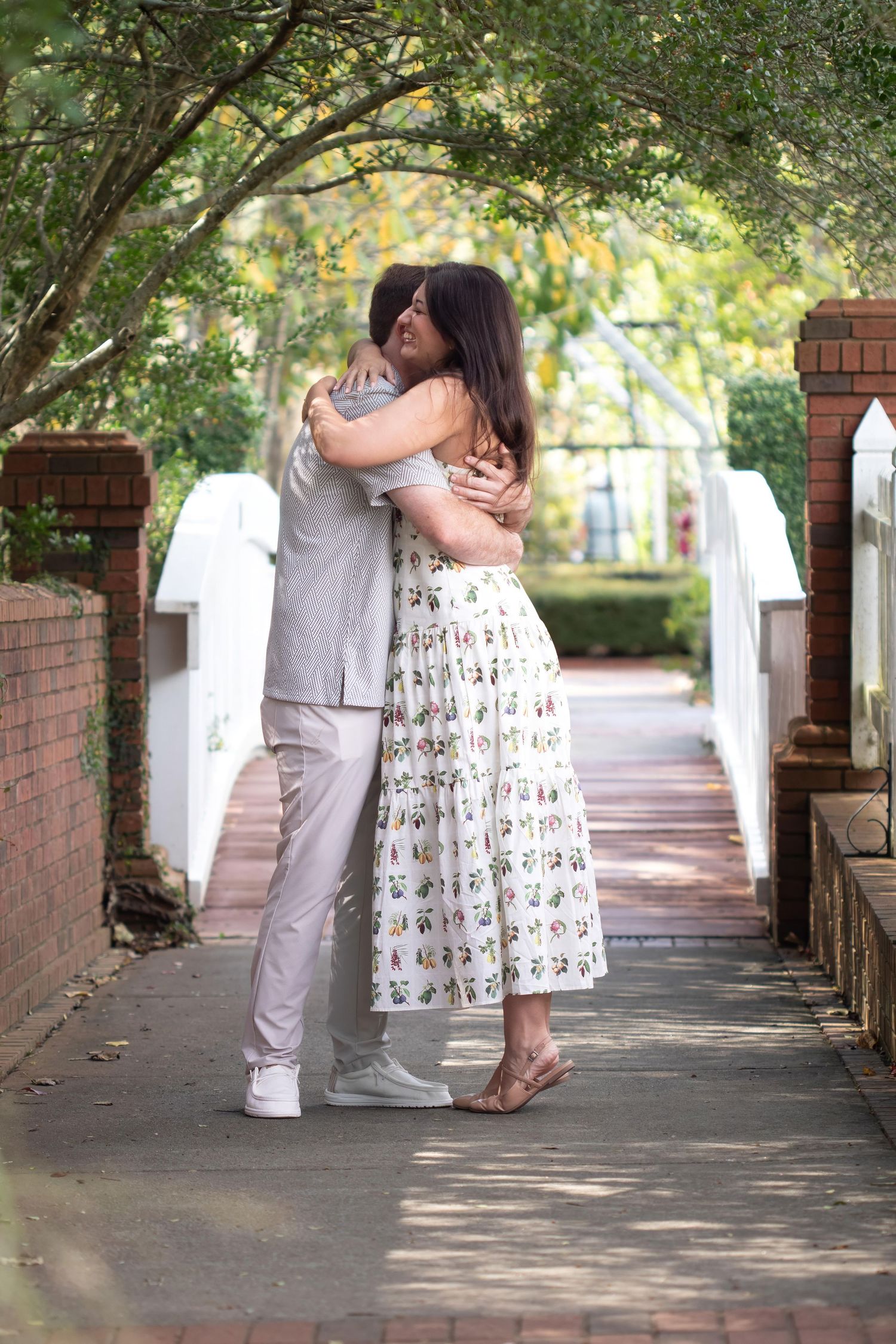 Couple embracing in a romantic outdoor walkway surrounded by lush green trees and brick pillars.