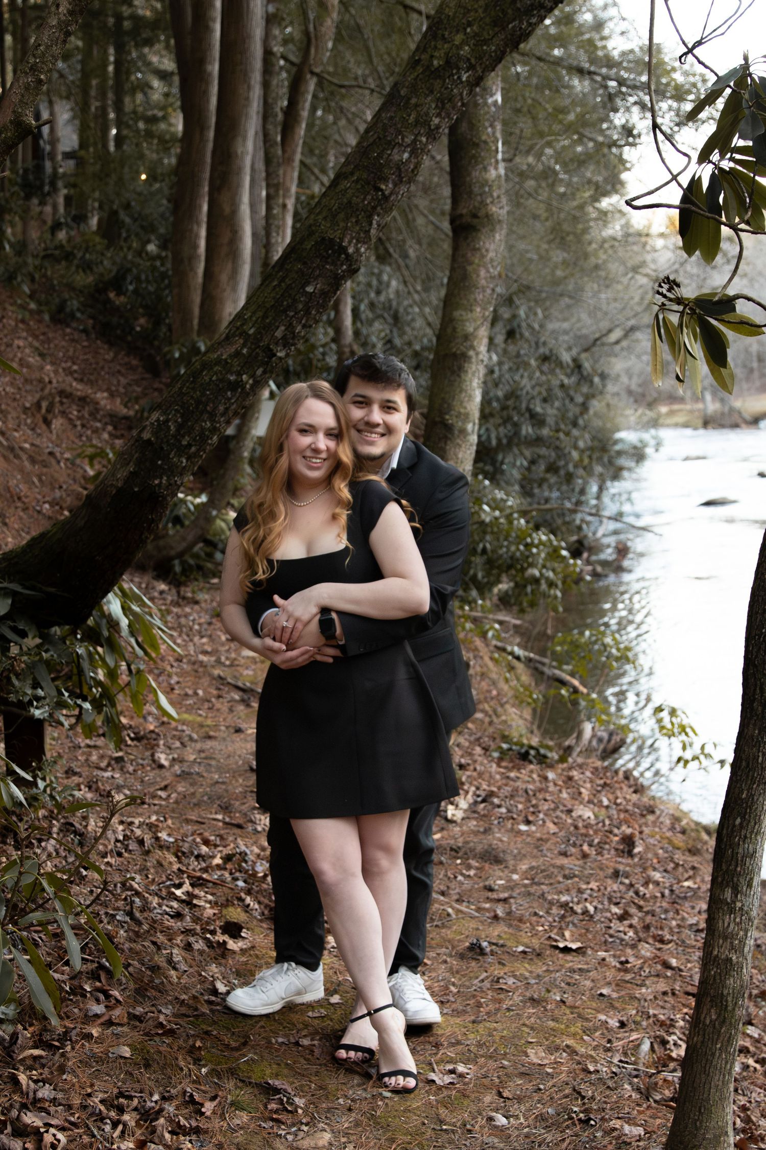A young couple in matching black formal attire poses embracing on a forest path beside a river.