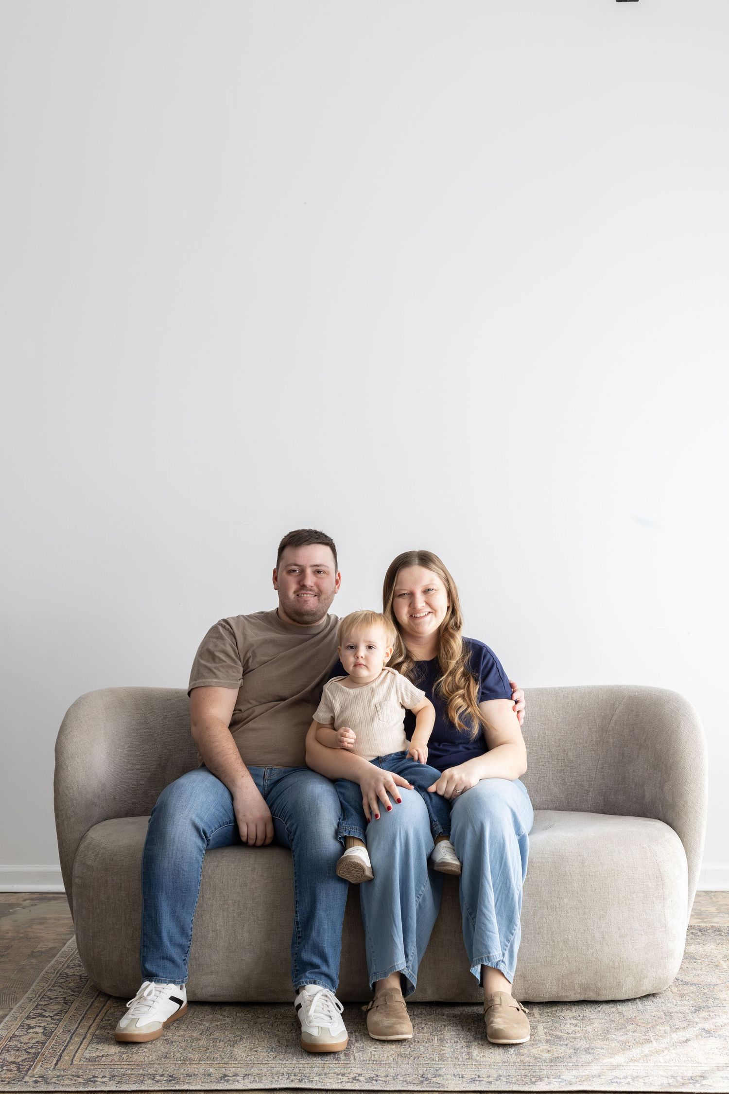 A young family with a baby sits smiling on a modern gray sofa against a white wall.