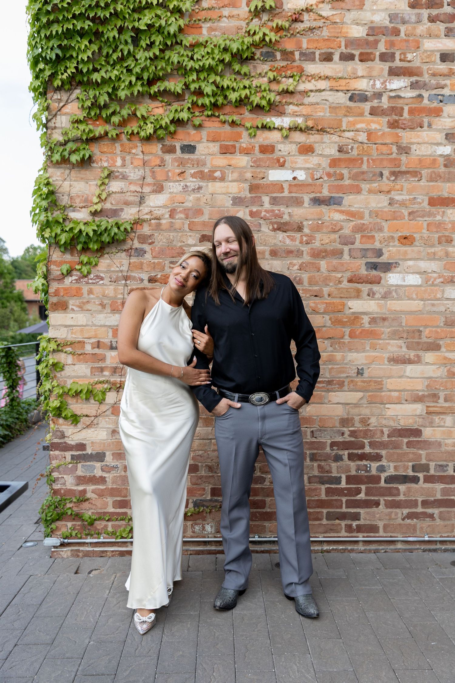 Couple posing against rustic brick wall with ivy, woman in white dress and man in black shirt and gray pants.