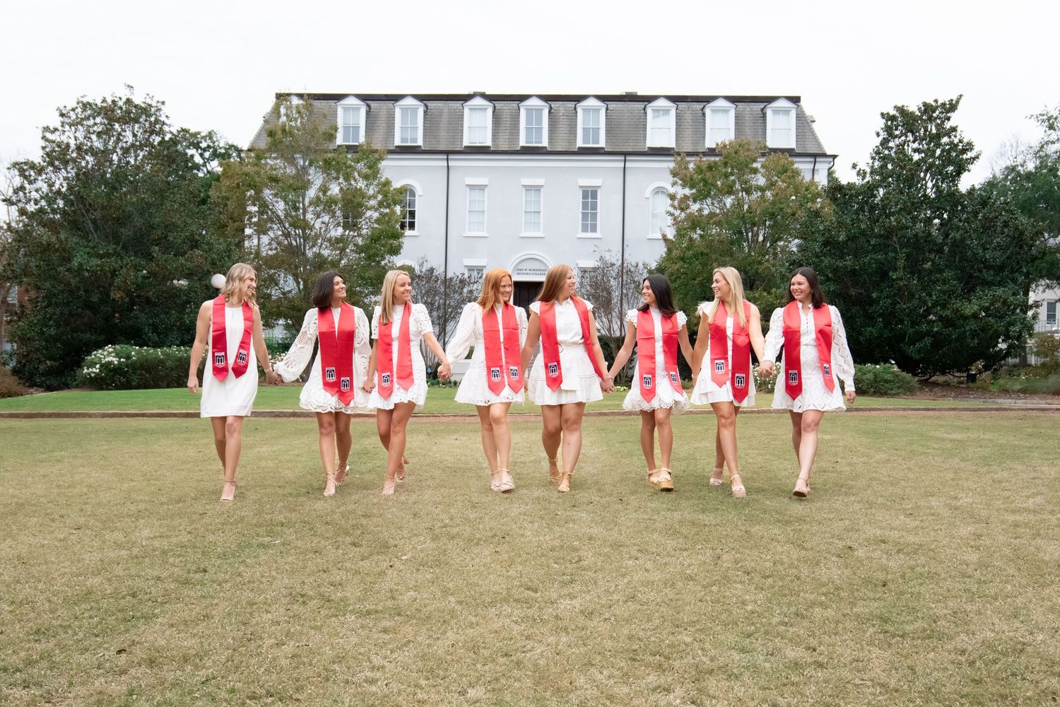 Seven women in white dresses and red sashes walk hand-in-hand on a green lawn in front of a large white building.