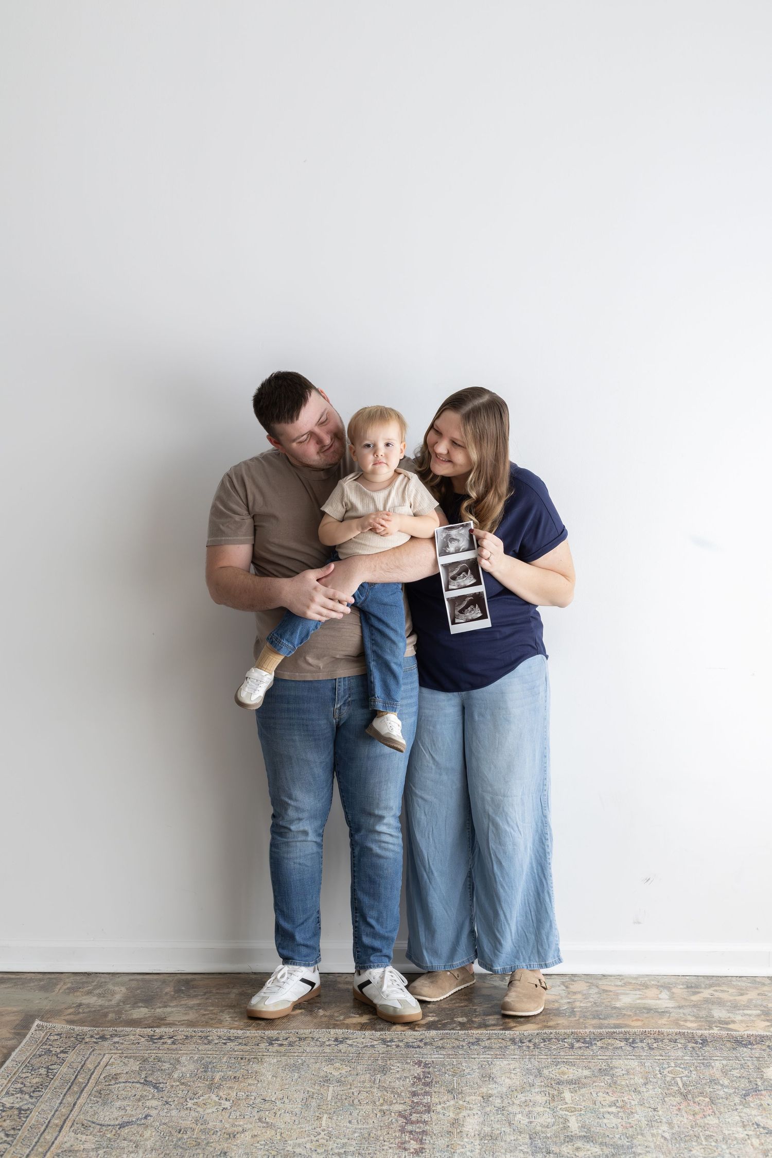 Happy couple holding toddler while woman displays ultrasound photo, announcing second pregnancy in bright studio.