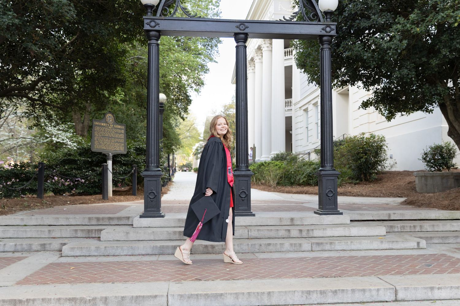 Woman in graduation gown walking in front of the UGA Arch on a sunny day.