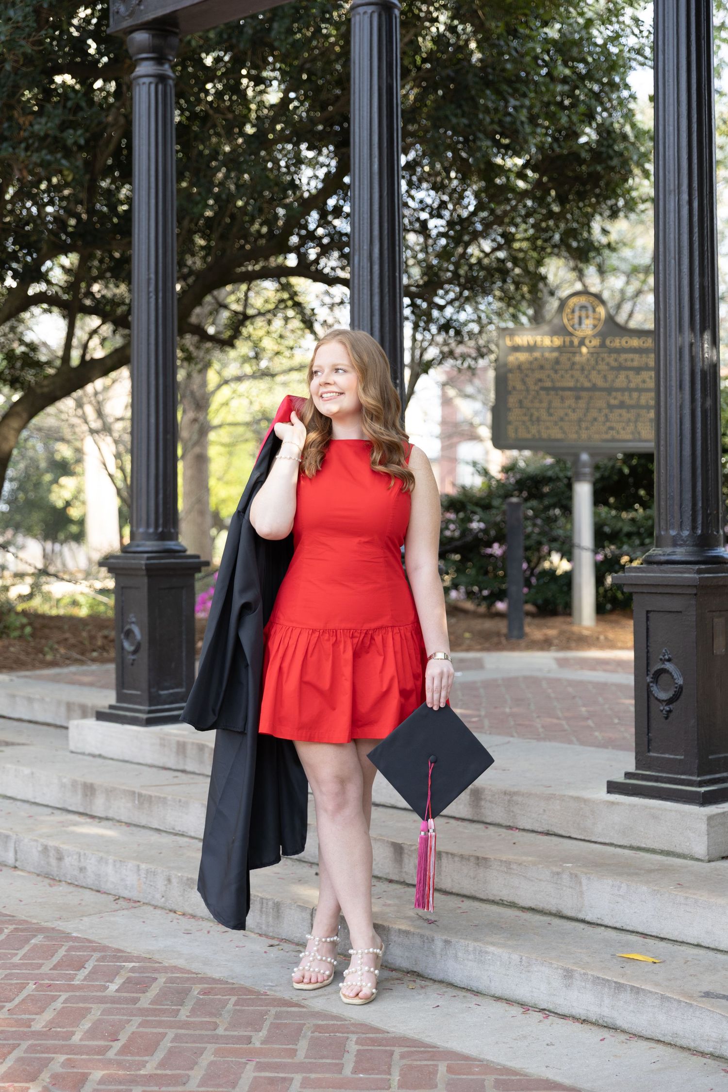Young woman in red dress holding graduation cap and gown, smiling outdoors near ornate black columns.