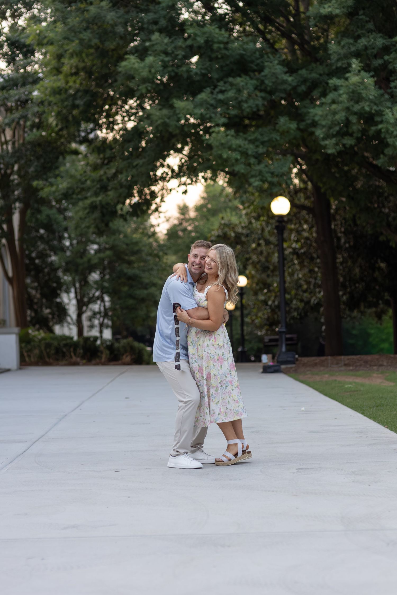 Happy couple embracing and dancing on outdoor plaza surrounded by lush green trees at sunset.