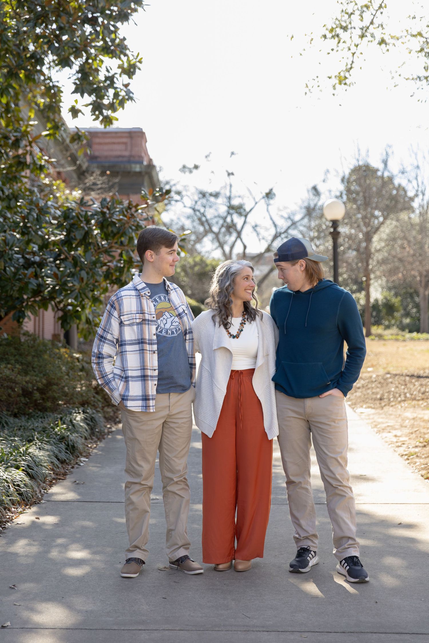 A woman in red pants and white cardigan stands smiling with two young men on a sunny campus walkway.