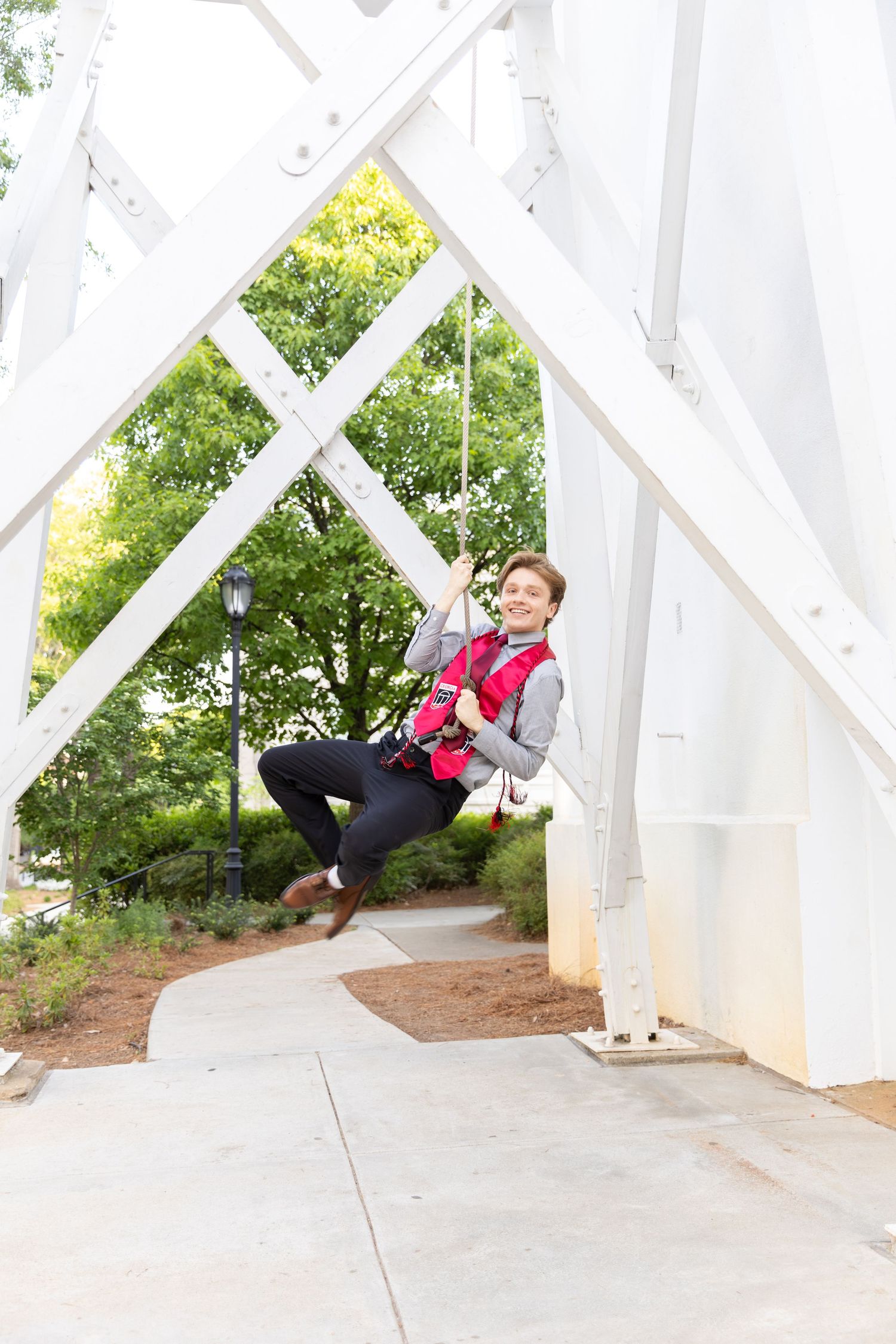 Smiling graduate in gray suit and red sash swings on a rope under a white bridge structure.