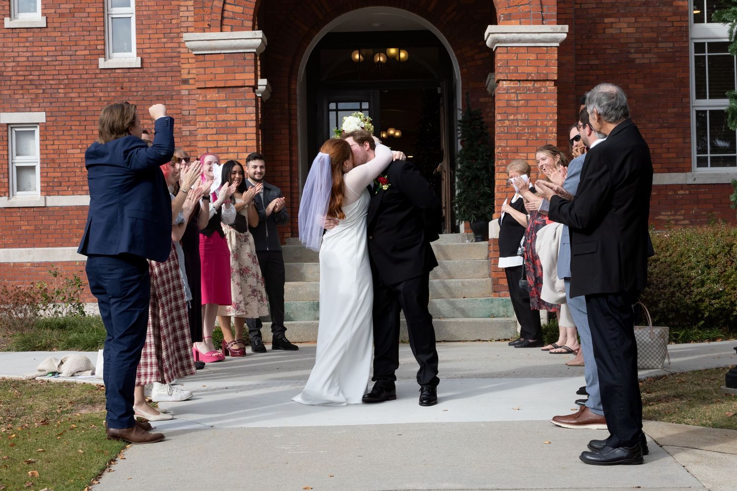 Newlyweds share a kiss outside a brick building as guests applaud and photograph the joyful wedding exit.