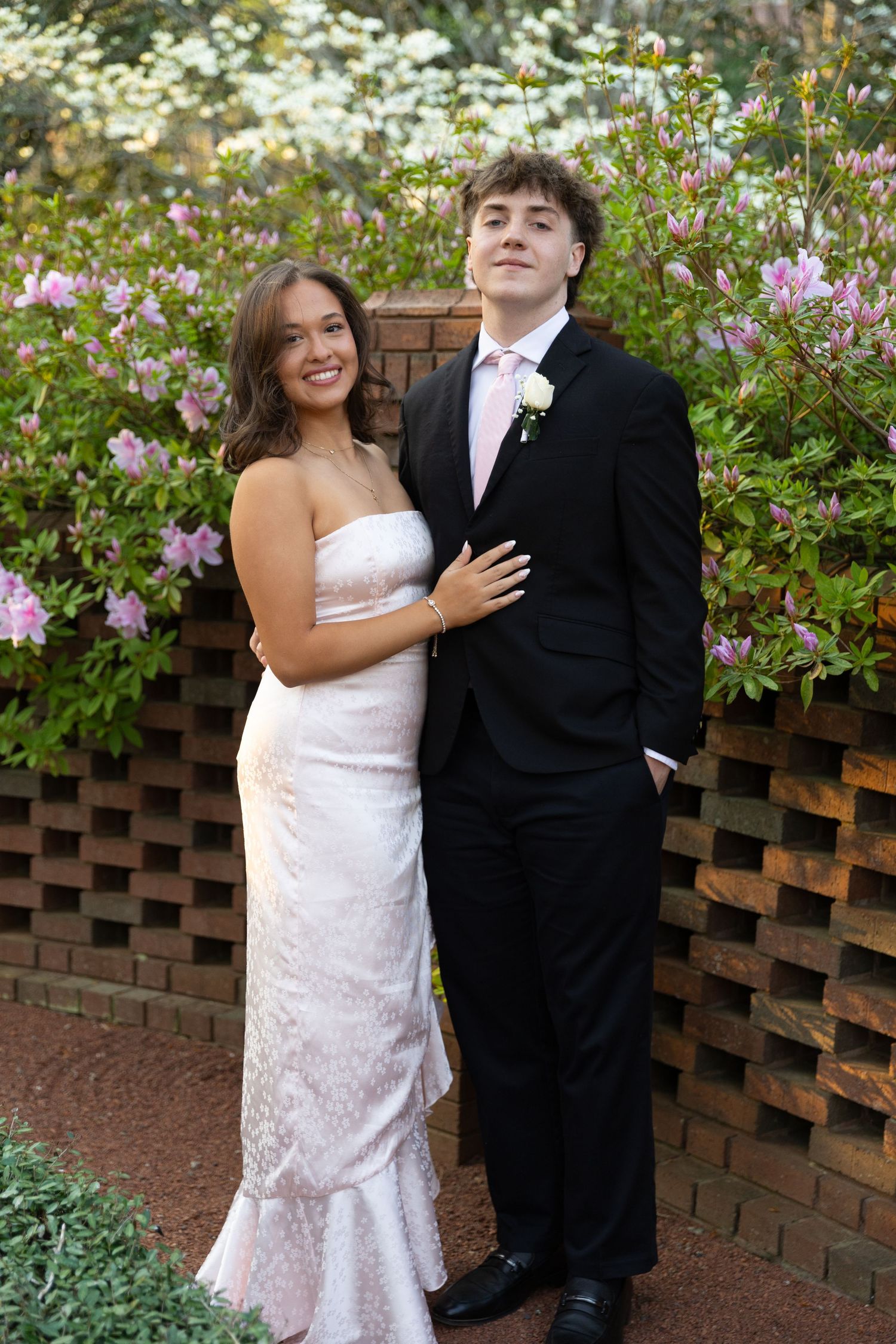 A young couple in prom attire poses outdoors, she in a white gown and he in a black tuxedo with pink tie.