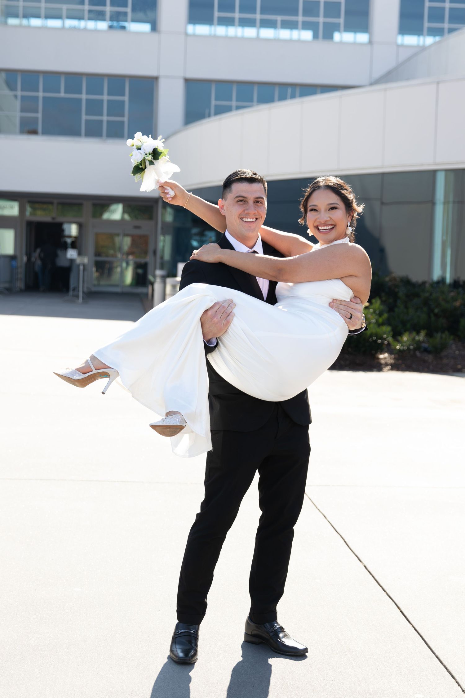 Smiling groom in black tuxedo carries bride in white wedding dress outside modern building on wedding day.
