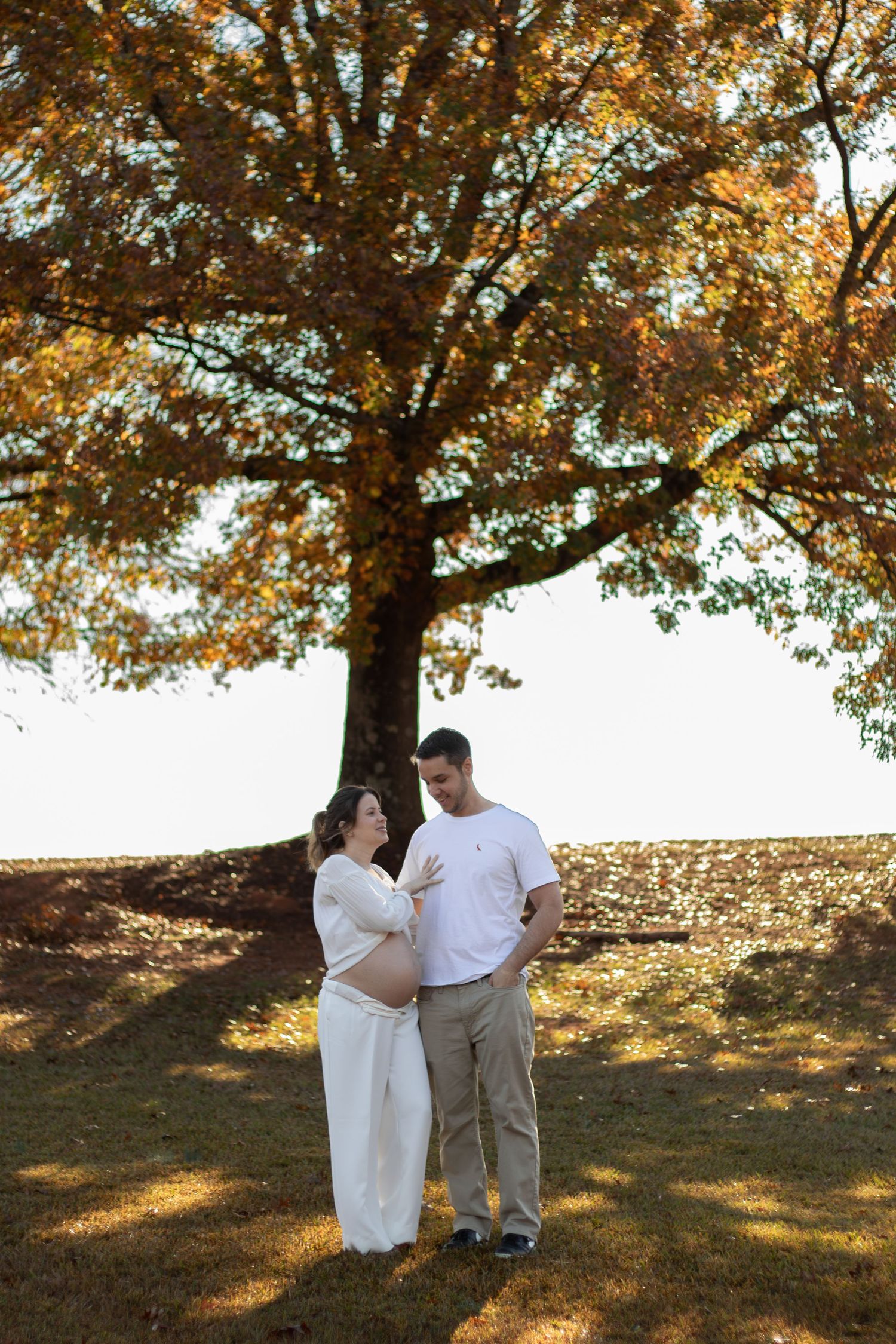 Expecting couple stands under autumn tree, woman in white dress cradling baby bump, man in white shirt and khakis.