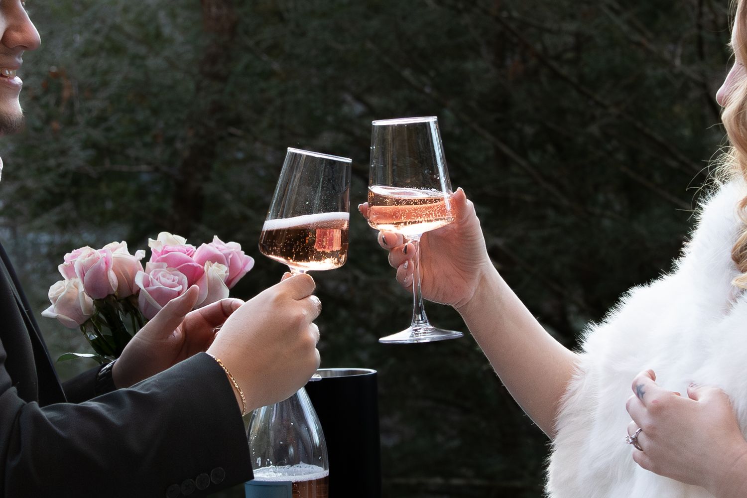 Couple clinking rose champagne glasses in a romantic outdoor toast, with pink flowers in the background.