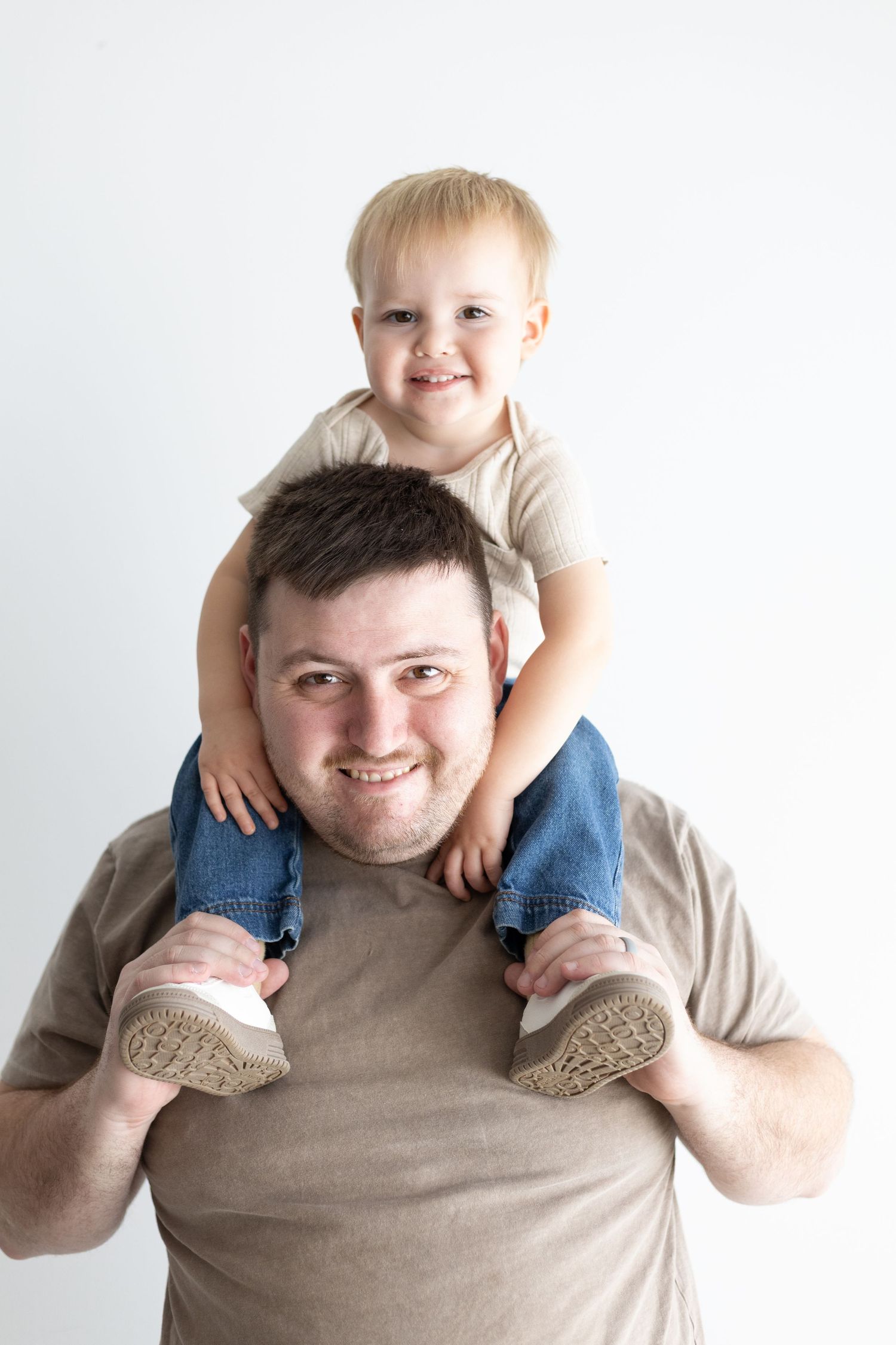 A smiling man carries a happy toddler boy on his shoulders against a white background.
