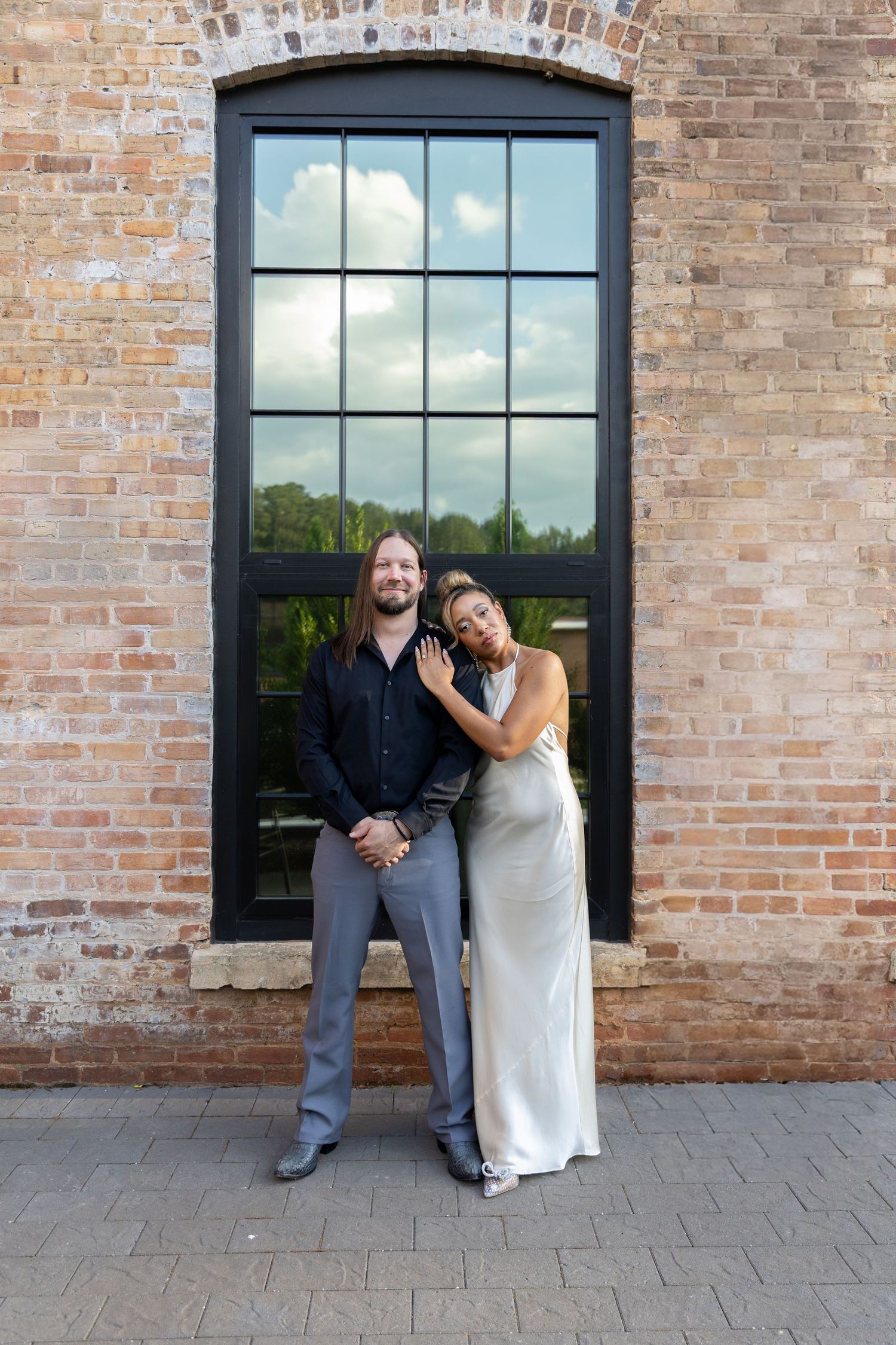 Couple posing in front of brick wall and large window, woman in silver gown leaning on man in black shirt.