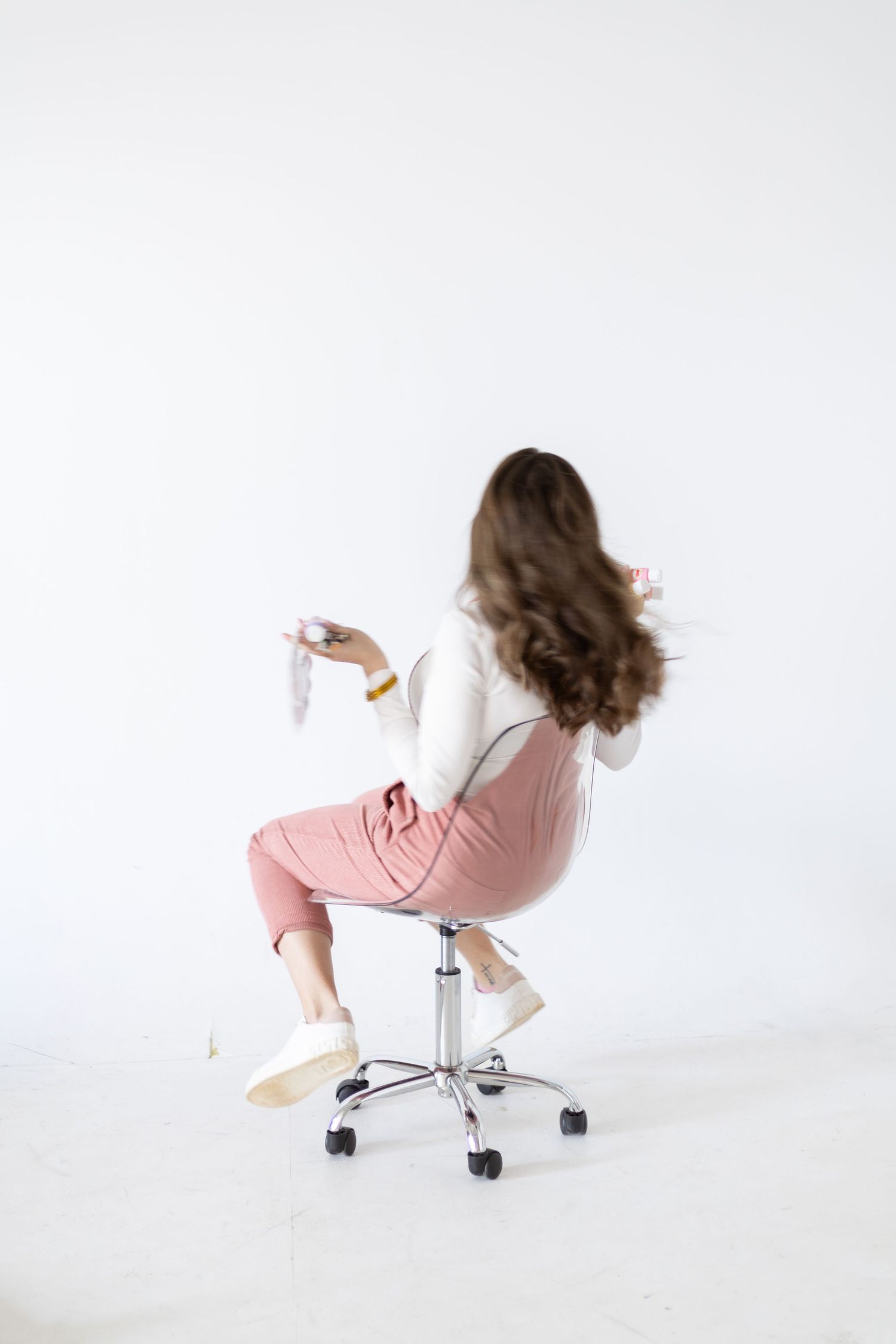 Young woman with curly hair sitting backwards on a rolling chair, wearing pink overalls and white top, holding objects.