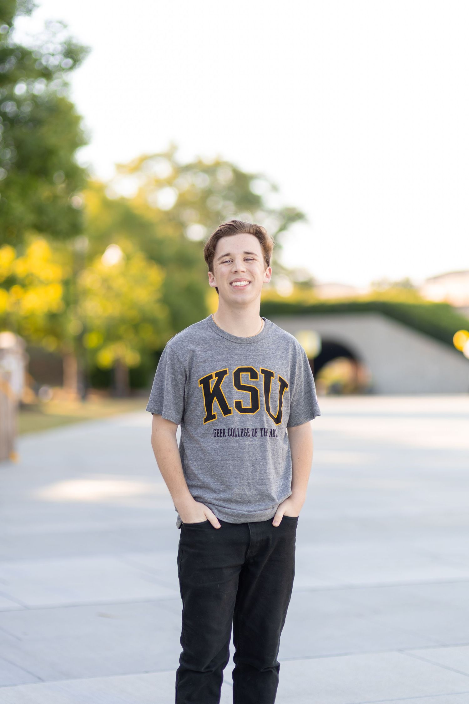 Young man smiling in a gray KSU college t-shirt and black jeans, standing outdoors on a campus pathway.