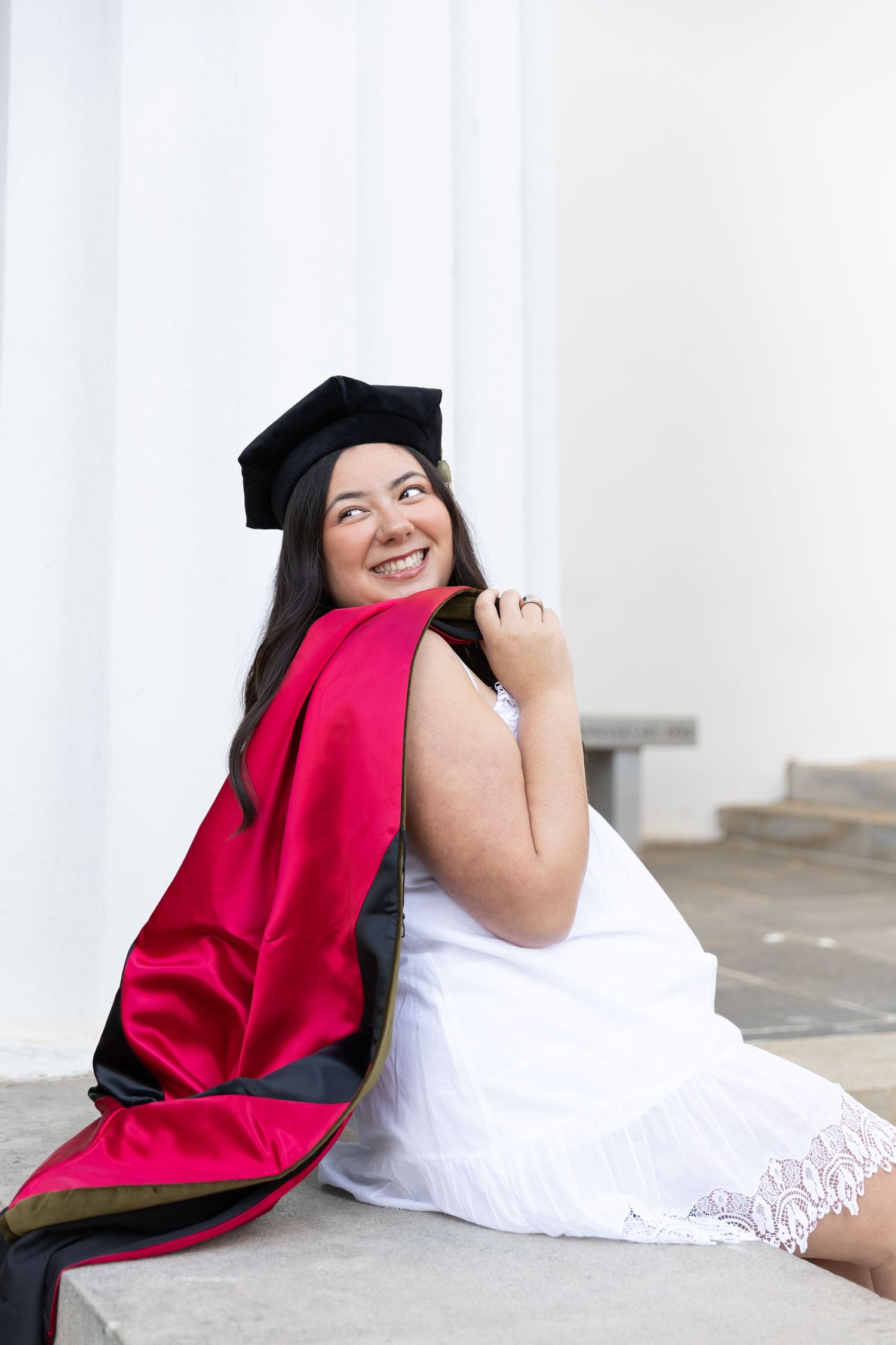 Smiling female graduate in black cap and red doctoral gown sitting on steps outdoors.