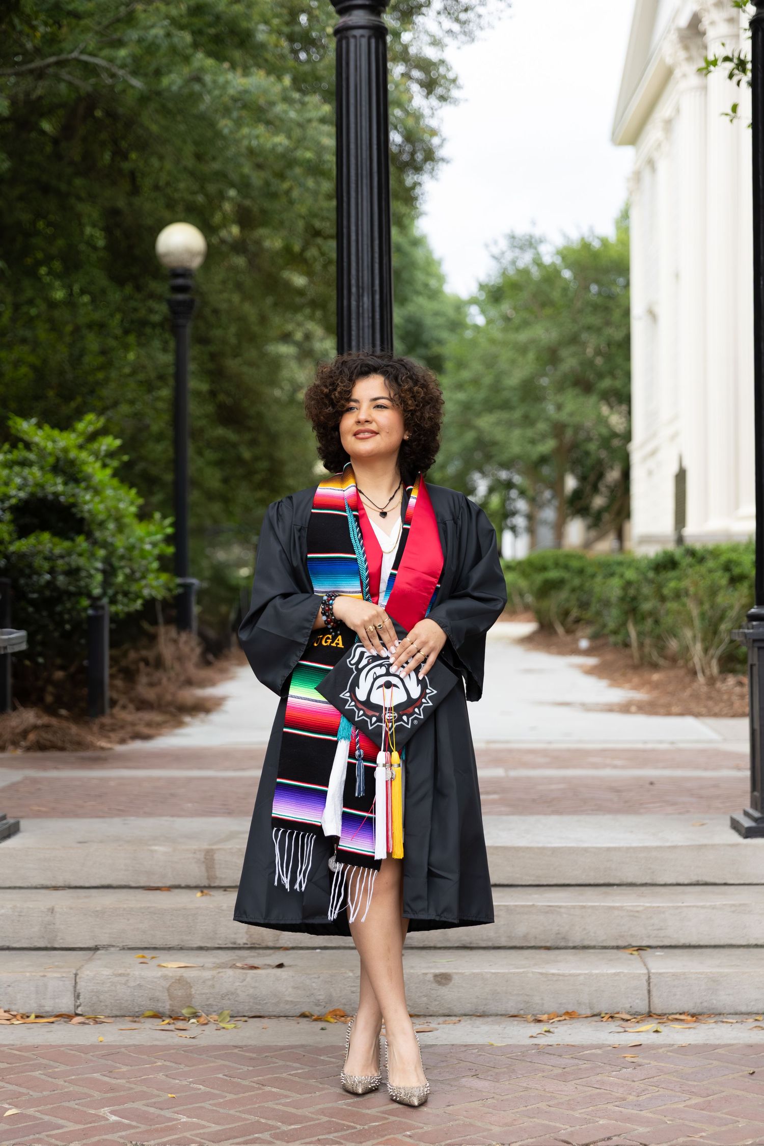 Young woman in graduation gown with colorful serape stole poses outdoors on campus steps holding decorative bag.