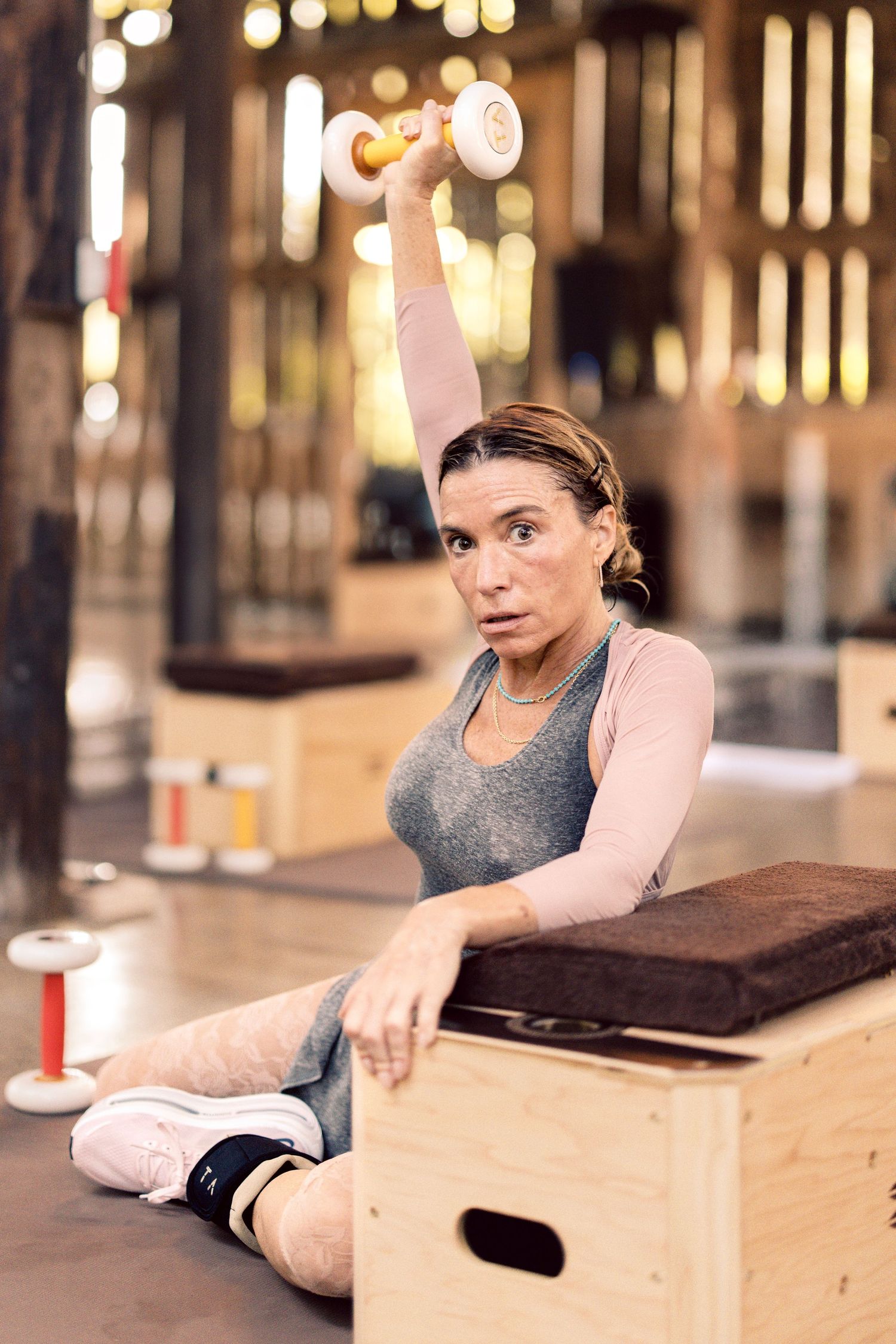 Woman sitting on wooden box in gym, lifting yellow dumbbell overhead during strength training workout.
