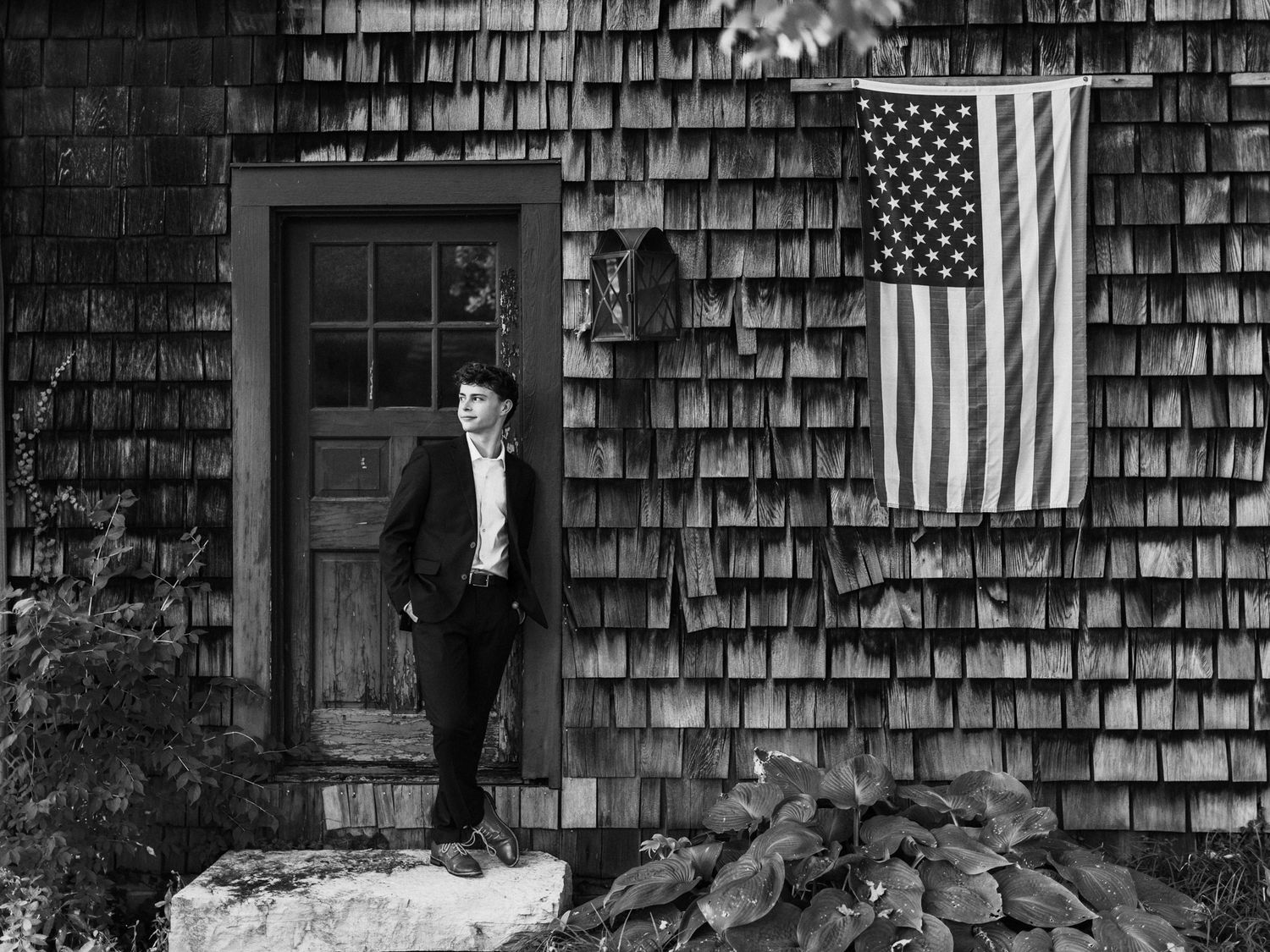 Black and white photo of a person standing beside a rustic shingle building with an American flag.