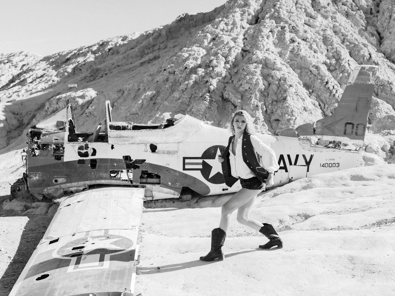 Black and white photo of a woman posing beside a vintage US Navy aircraft in a snowy mountain setting.