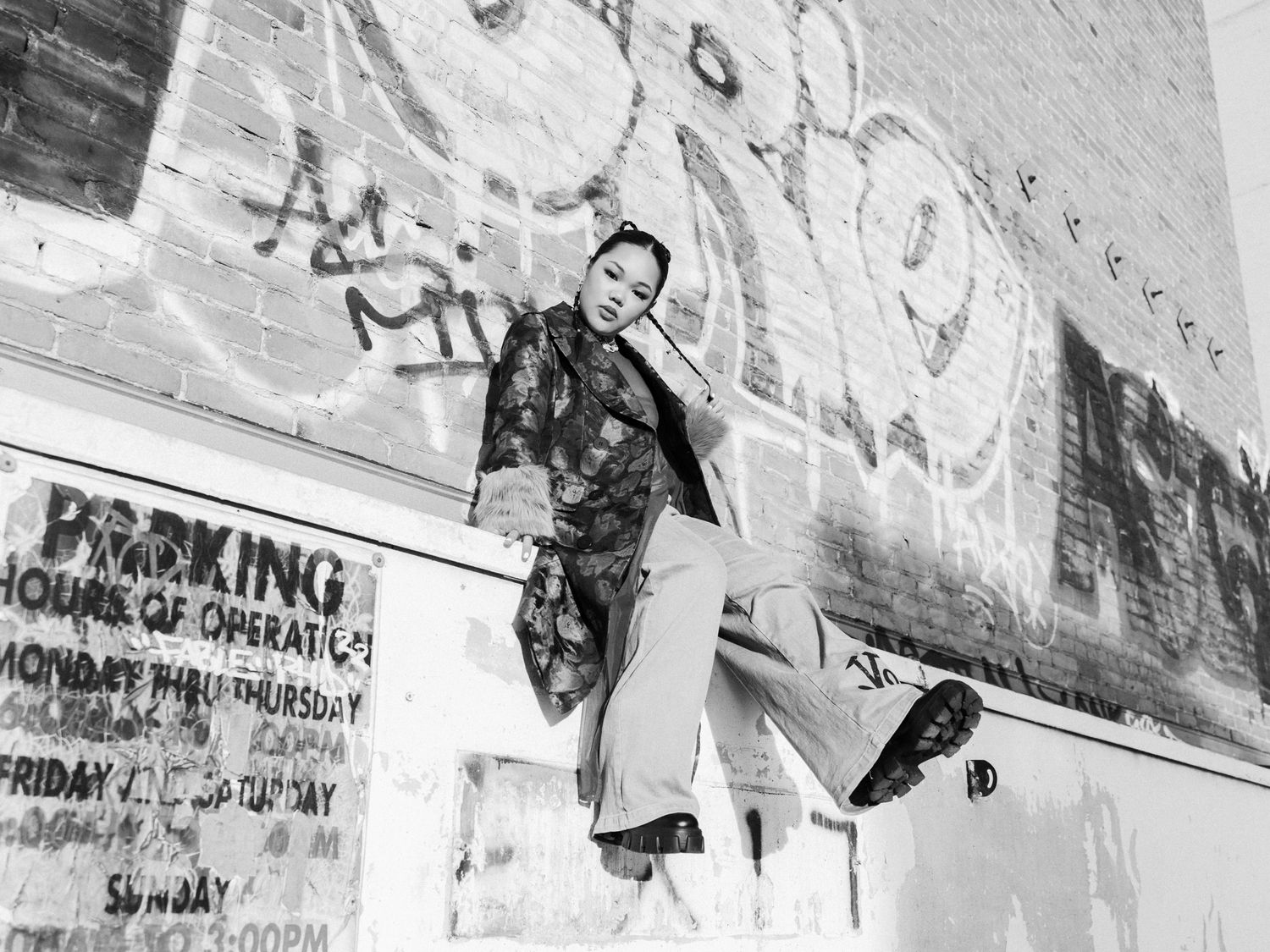 Black and white photo of woman in patterned jacket posing against graffiti wall near parking sign.